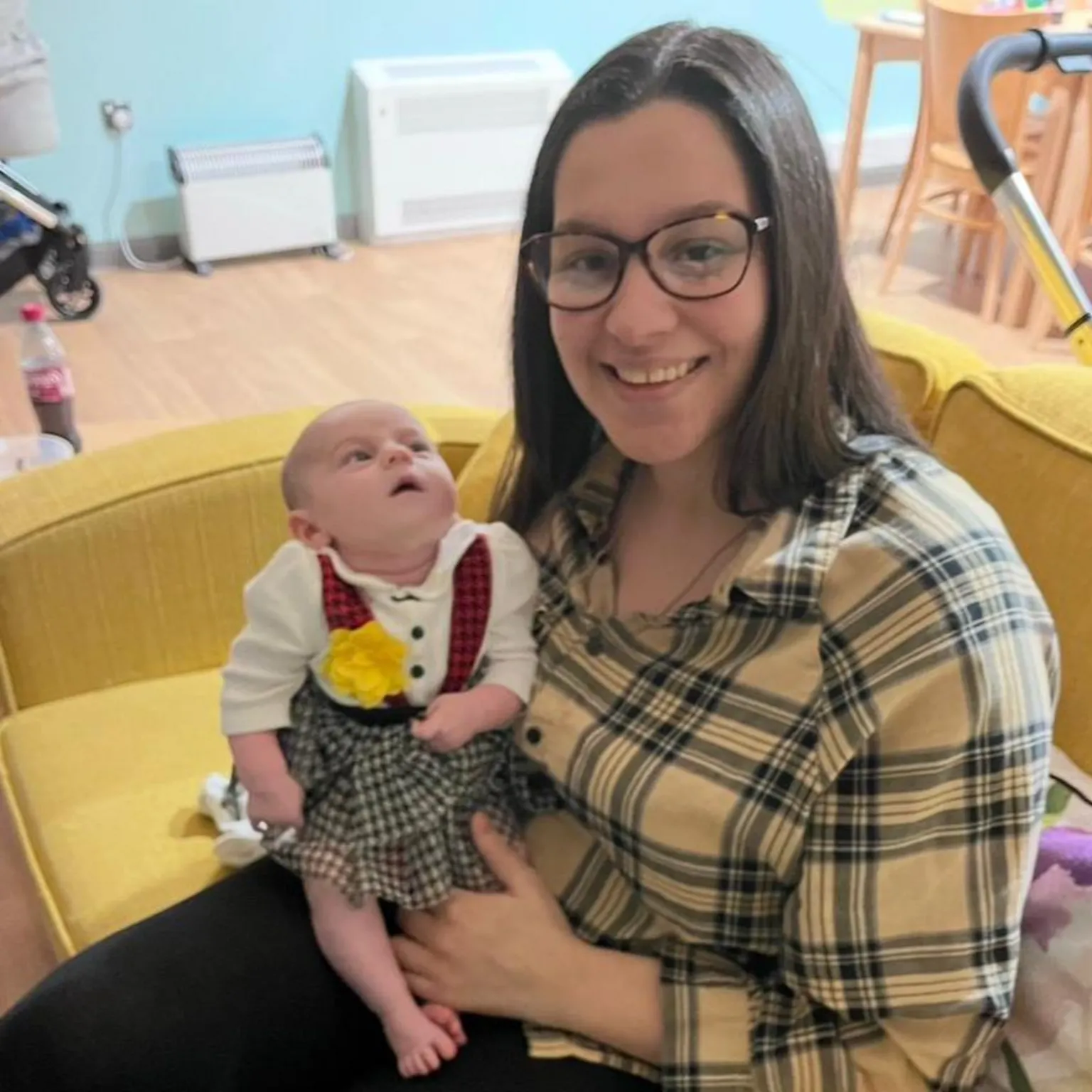 Sofii Lewis A woman smiles at the camera. She has shoulder-length brown hair and glasses on. She wears a yellow checked shirt and black trousers and is sat on a yellow sofa. She hold her baby who is in a Welsh lady outfit.