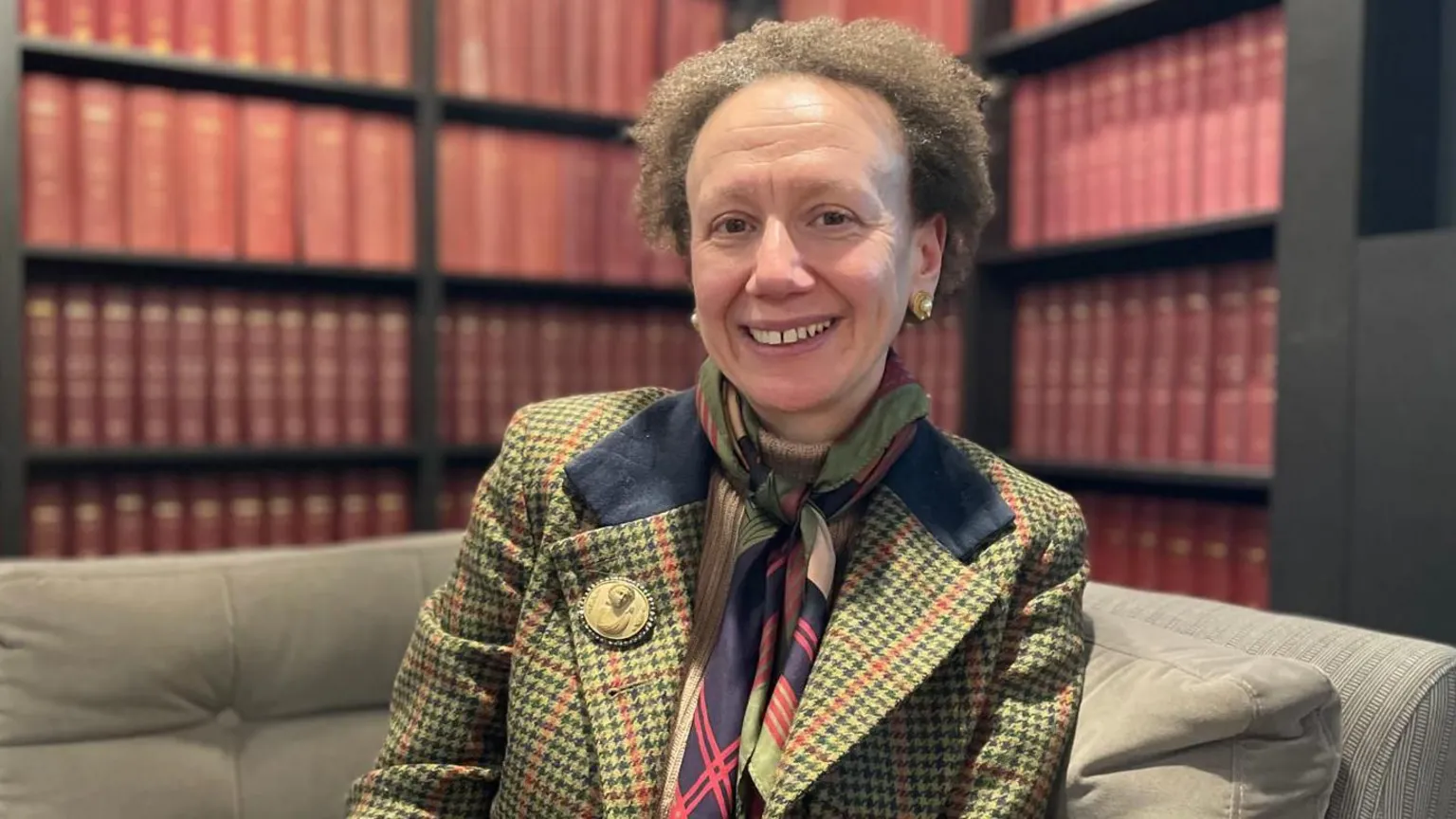 A woman in a green, navy and red checked jacket, with a similarly patterned scarf smiles at the camera. She has short, brown and curly hair and is sat on a grey sofa in front of bookcases filled with red, leather bound books.