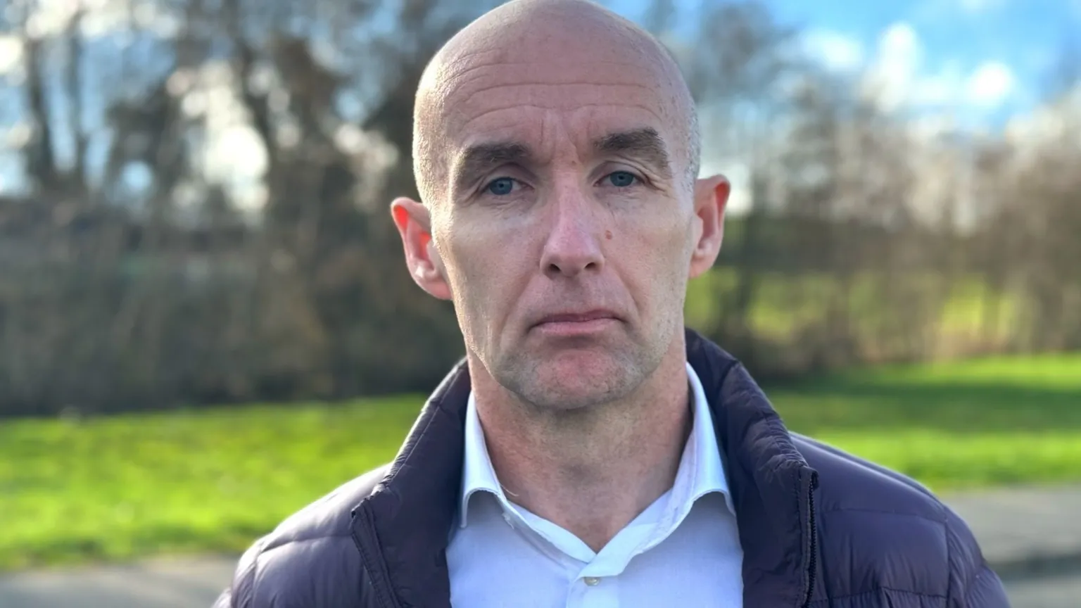 A man, who is bald, wearing a white shirt and black puffer jacket.