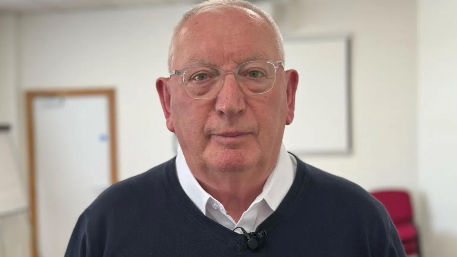 An older man who is partially bald with short hair wearing silver glasses. He has a white shirt on and navy v-neck jumper.