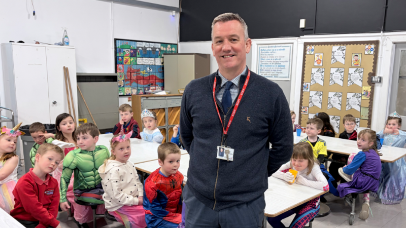 Dyn yn sefyll mewn ffreutur ysgol yn gwisgo siwmper las dros grys a thei tywyll a phlant mewn gwisg ffansi yn estedd o gwmpas byrddau tu ol iddo.