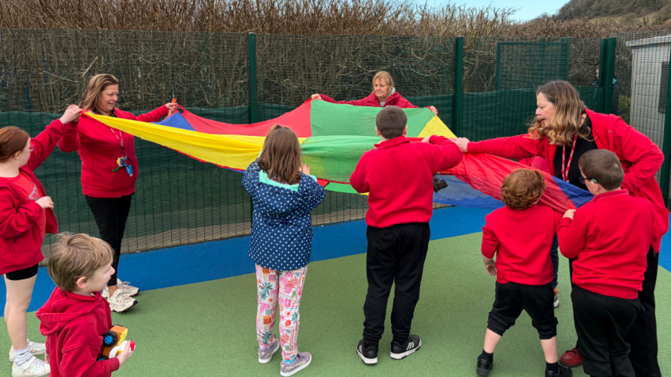 Grwp o blant mewn siwmperi coch ar iard ysgol yn dal canopi parachute lliwgar gydag aelodau staff yn helpu