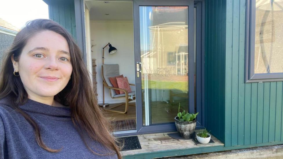 Llun o Elen Rennolf y tu allan i'w swyddfa gwnsela sydd wedi ei gosod yn yr ardd. Mae hi'n dal y camera ac yn gwenu. Mae ganddi wallt hir brown.