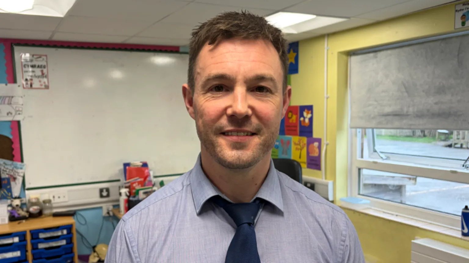 A man in a classroom with short brown hair wearing a light blue shirt and navy tie