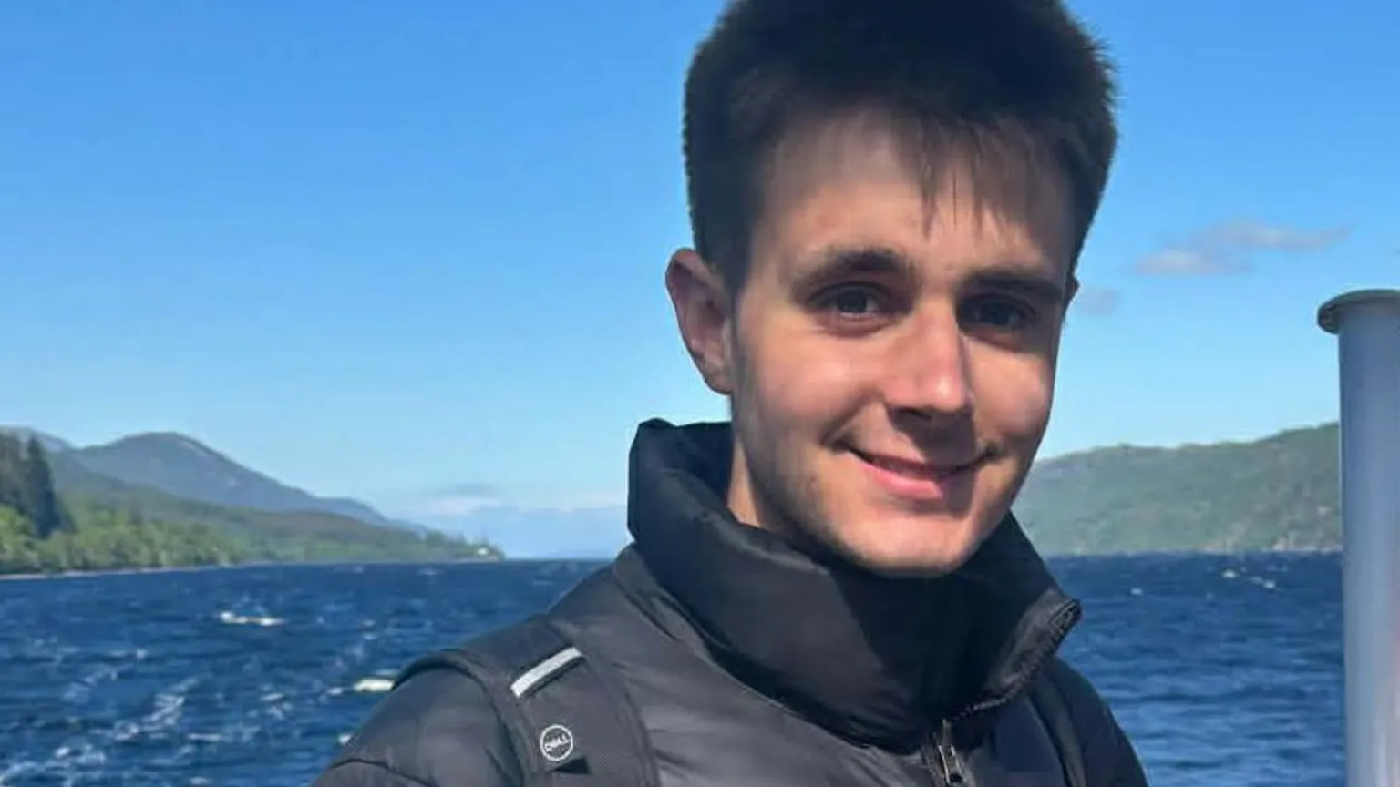 Supplied A head and shoulders image of James Thompson. A young man wearing a black puffer jacket with two black rucksack straps over it. He appears to be on a boat, which has Union Jack flag on it. There is water and mountains in the background.