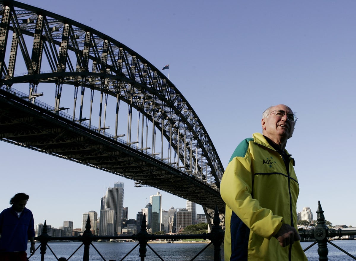 John Howard wears a yellow and green ‘Australia’ tracksuit top with the Sydney Harbour Bridge in the background
