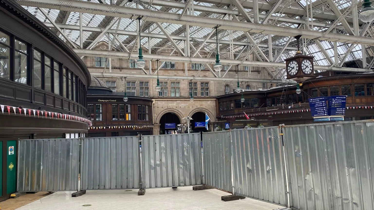 The main waiting area of Glasgow Central by the Gordon Street entrance. The waiting area is obscured by metal sheeting and there is a glass roof held up by white barring. The place is empty