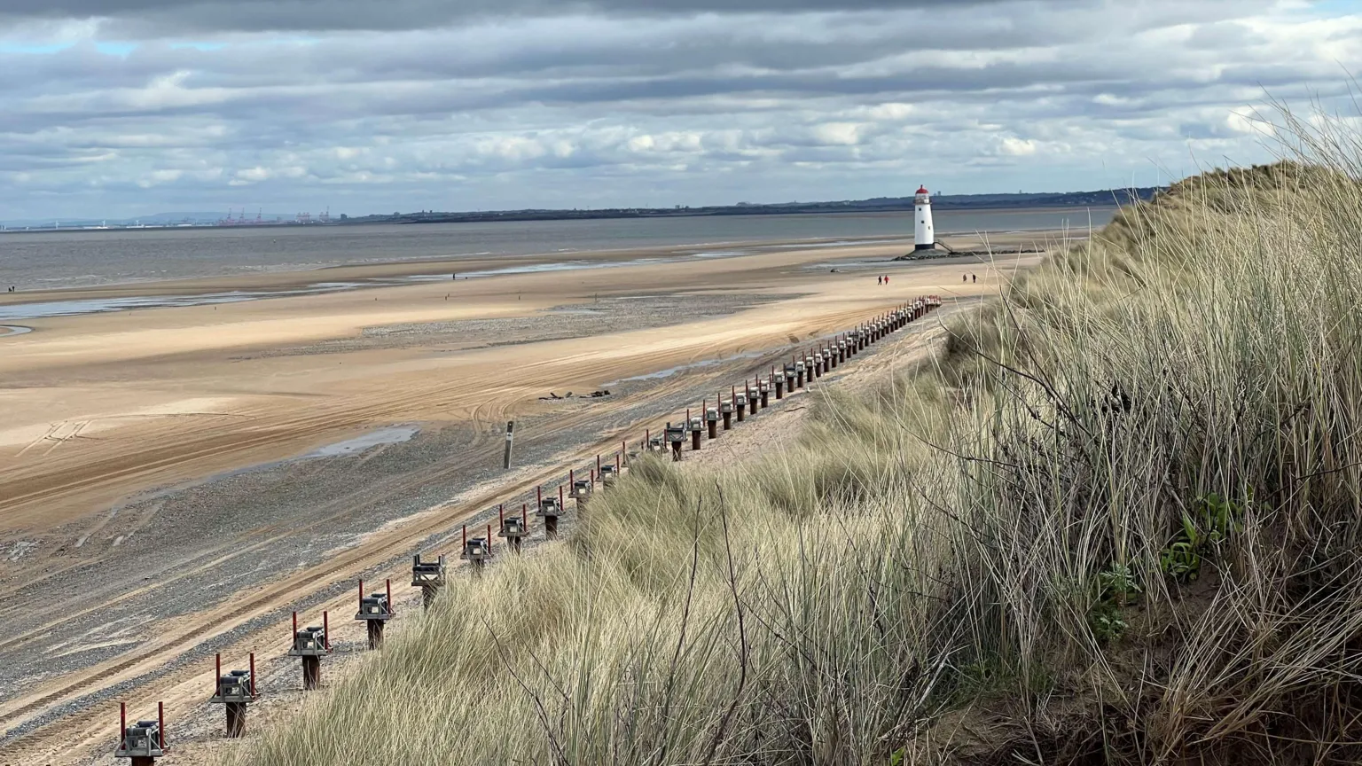 Eni This image shows Talacre Beach, the sky is blue and there's a lighthouse in the background, you can see the line of structures on the beach. There's grass in the foreground.