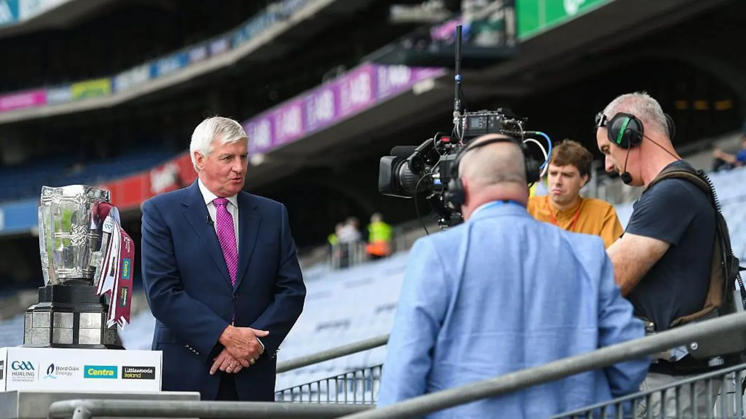 A short-grey haired man wears a navy suit, white shirt and pink tie. He speaks into a black TV camera, which is being held by a shirt-grey haired man in a navy t-shirt, wearing a headset. Beside the man in the navy suit is a silver trophy with white and maroon ribbons wrapped around its handle. A bald man in a blue suit and a short-brown haired man in an orange shirt look on from behind the camera. There are rows of empty blue seats behind them all. 