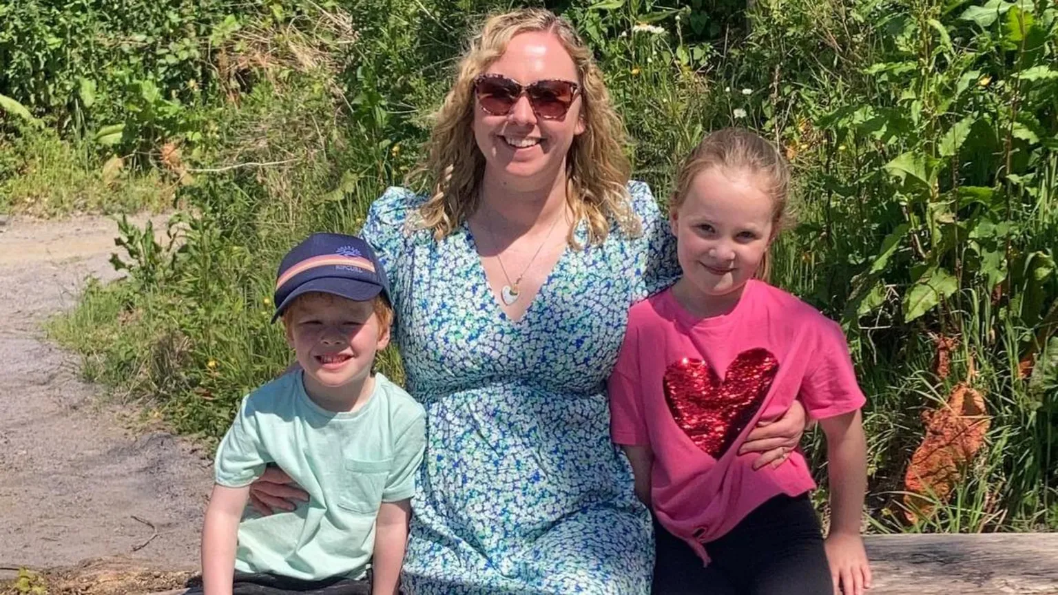 Fflur Evans Fflur Evans, wearing sunglasses and a blue floral summer dress, with two her children. Dafi, on the left, is wearing a cap and blue t-shirt. Anni, on the right, has her hair tied back and she's wearing a pink t-shirt with a red heart. The three are smiling as they sit on a bench in a park. There are bushes behind them.
