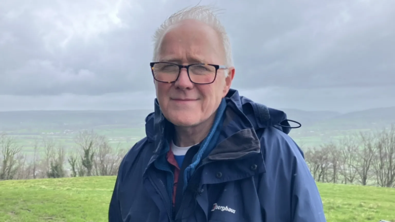 Rob Messenger has short white hair and black glasses. He is wearing a navy blue rain coat and is pictured near Paxton's Tower in Carmarthenshire. The sky is cloudy.