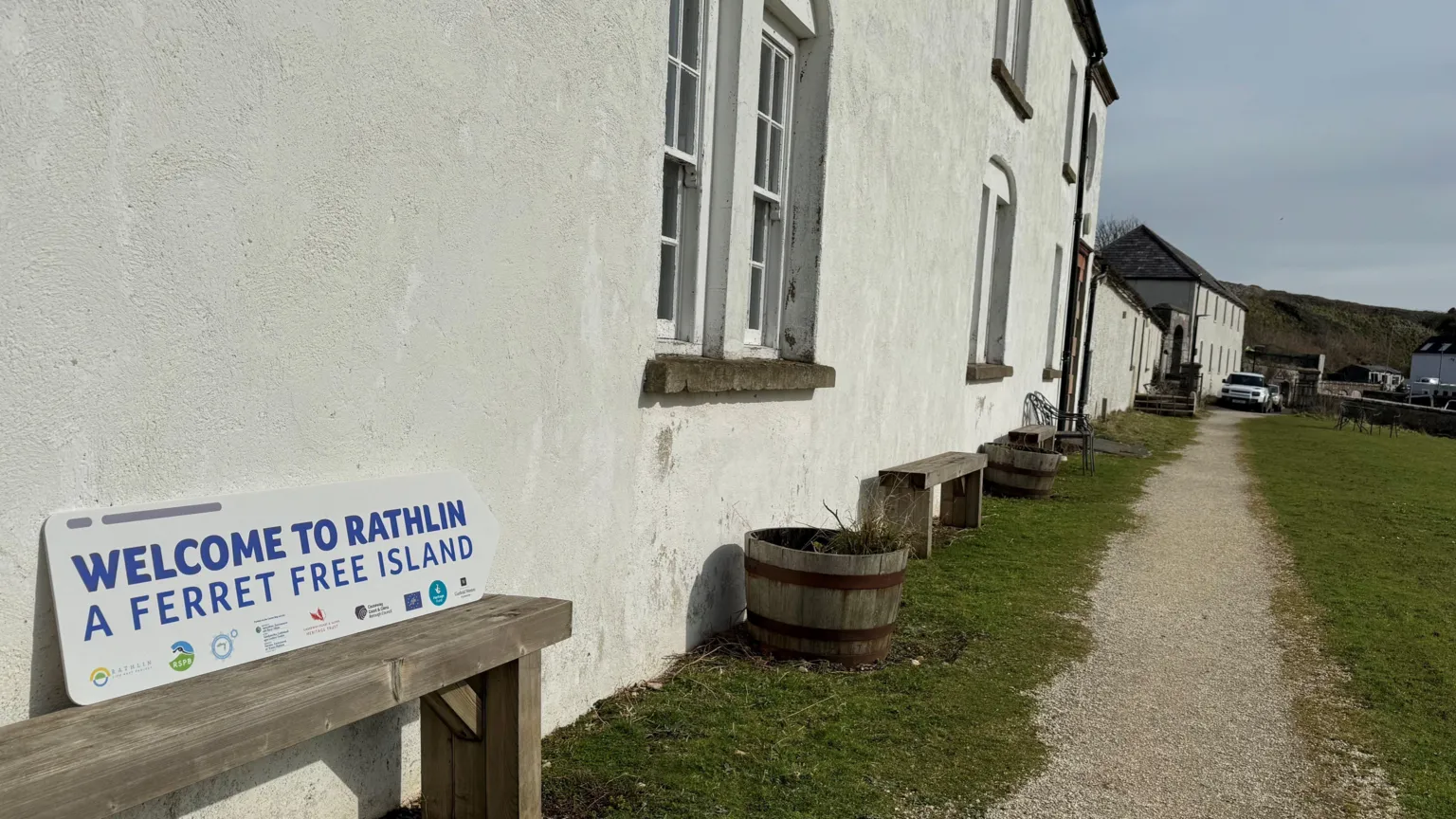 A sign says welcome to Rathlin, a ferret free island. It's been placed on a wooden bench next to a house. 