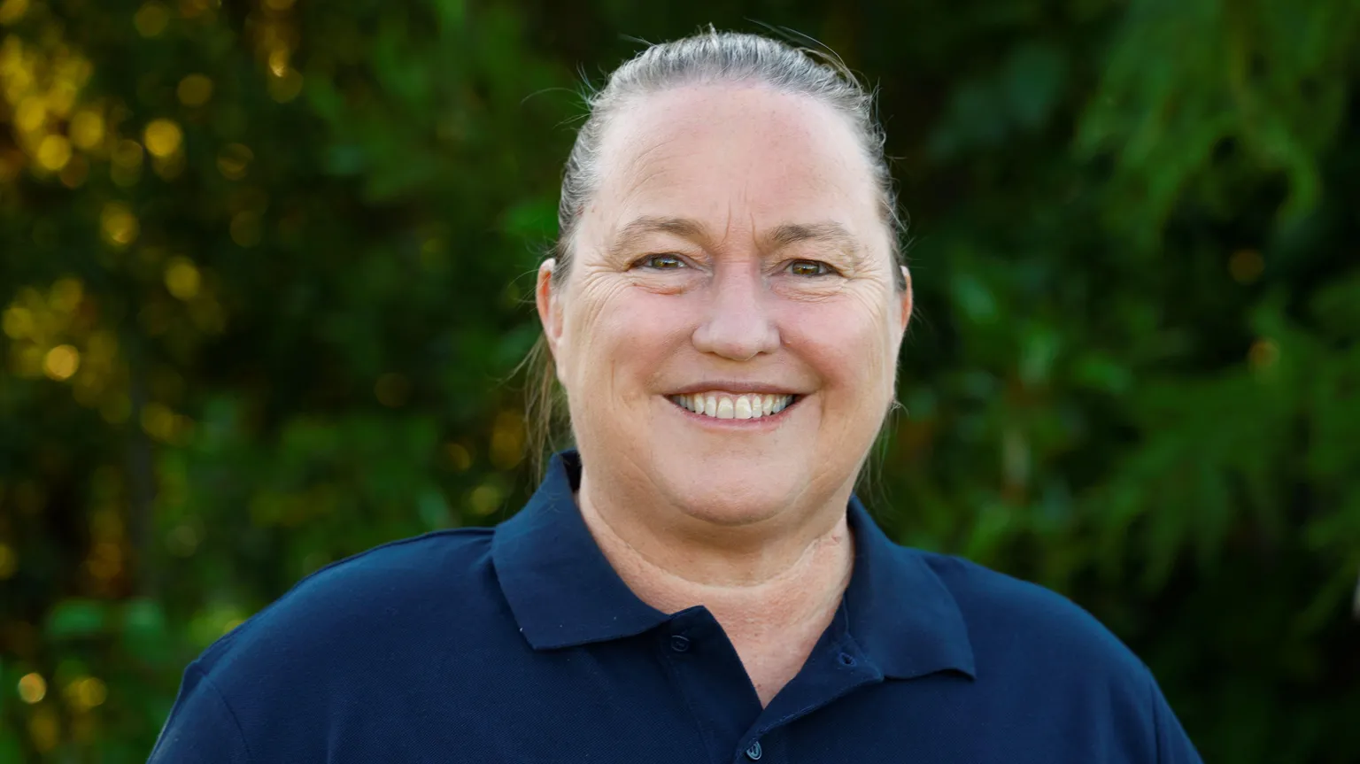 Phelps Visuals Biz is wearing a navy t-shirt. Foliage is out of focus in the background. 