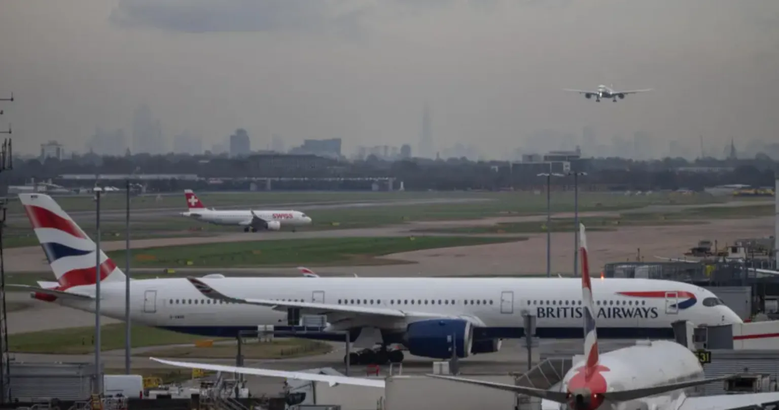 Planes arriving and departing from the runway at London Heathrow. One plane is coming into land, whilst others traverse the runways on the ground, where a BA plane parks at a gate whilst another smaller Swiss Airline plane also taxis behind it. The London skyline is clearly visible in the distance on a grey cloudy day, with the Shard being especially prominent.