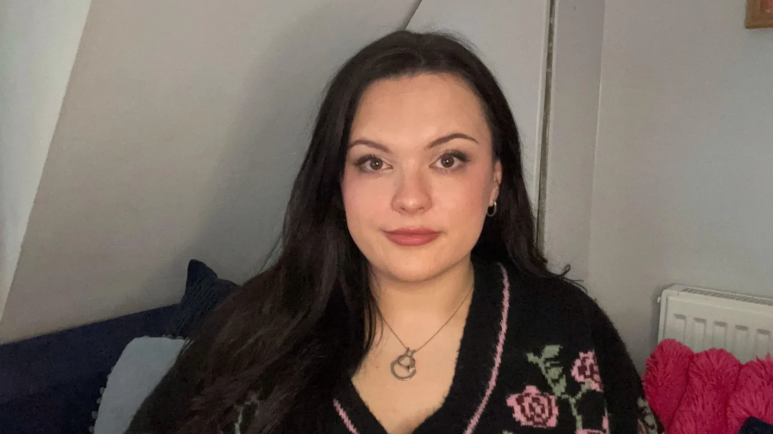 Jessica smith is a young woman. She has pale skin and long black hair that is slightly wavey. She has rosy cheeks and lips and is looking directly at the camera. She is wearing a fluffy cardigan with flowers on it, and a silver necklace with small silver hoop earrings. 