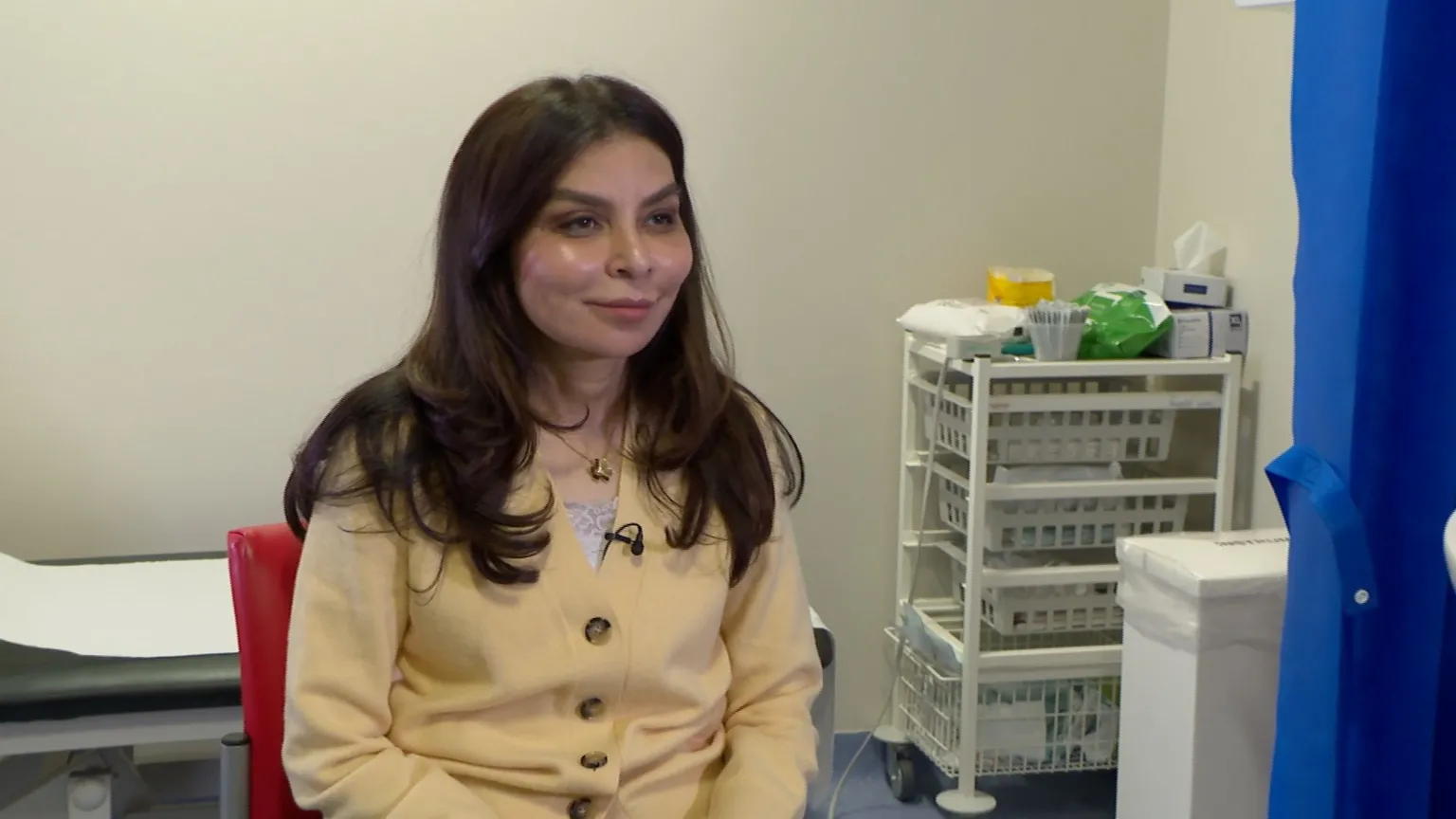 Consultant Gaity Ahmad is sat in her office, there is an examination bed in the background and a tray of needles, equipment and test tubes. She is wearing a yellow cardigan that is buttoned up and a gold necklace. She has shoulder-length, layered brown hair and is smiling. 