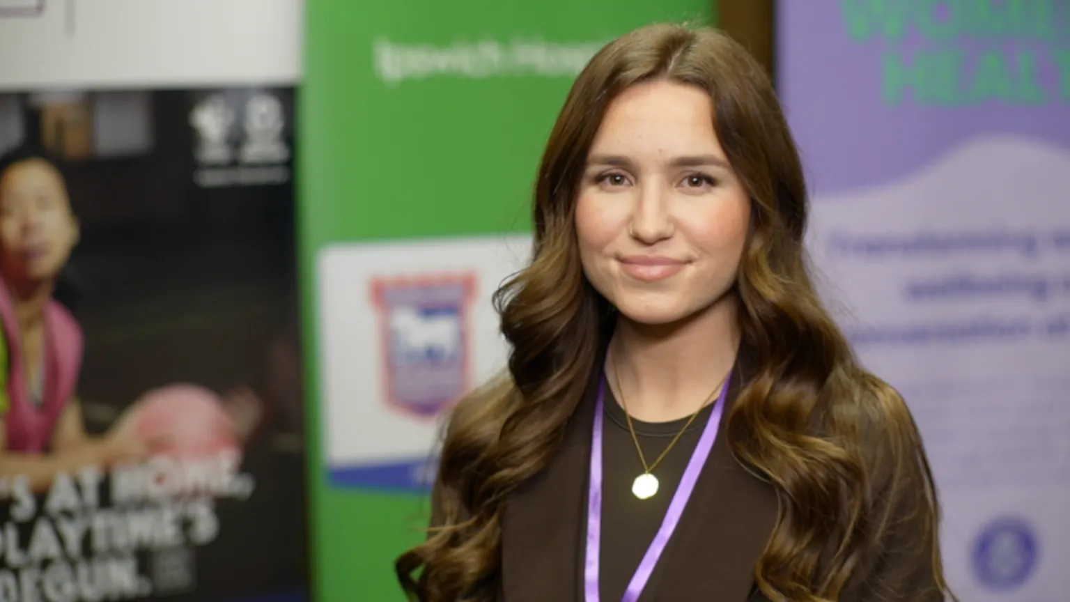Jamie Niblock/BBC Amy Peckham-Driver standing in front of information boards. She is looking directly at the camera and smiling and is wearing a brown T-shirt and purple lanyard.