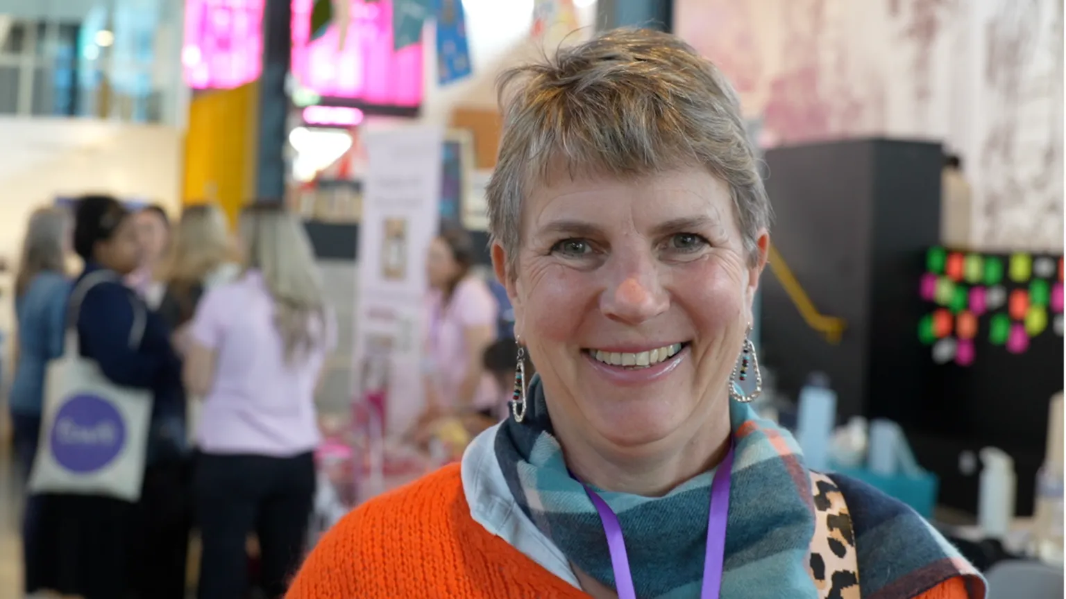 Jamie Niblock/BBC Julia Endacott, a woman standing inside a hall. She has short hair and is looking directly at the camera and smiling. She is wearing an orange jumper with a blue scarf and dangly earrings.