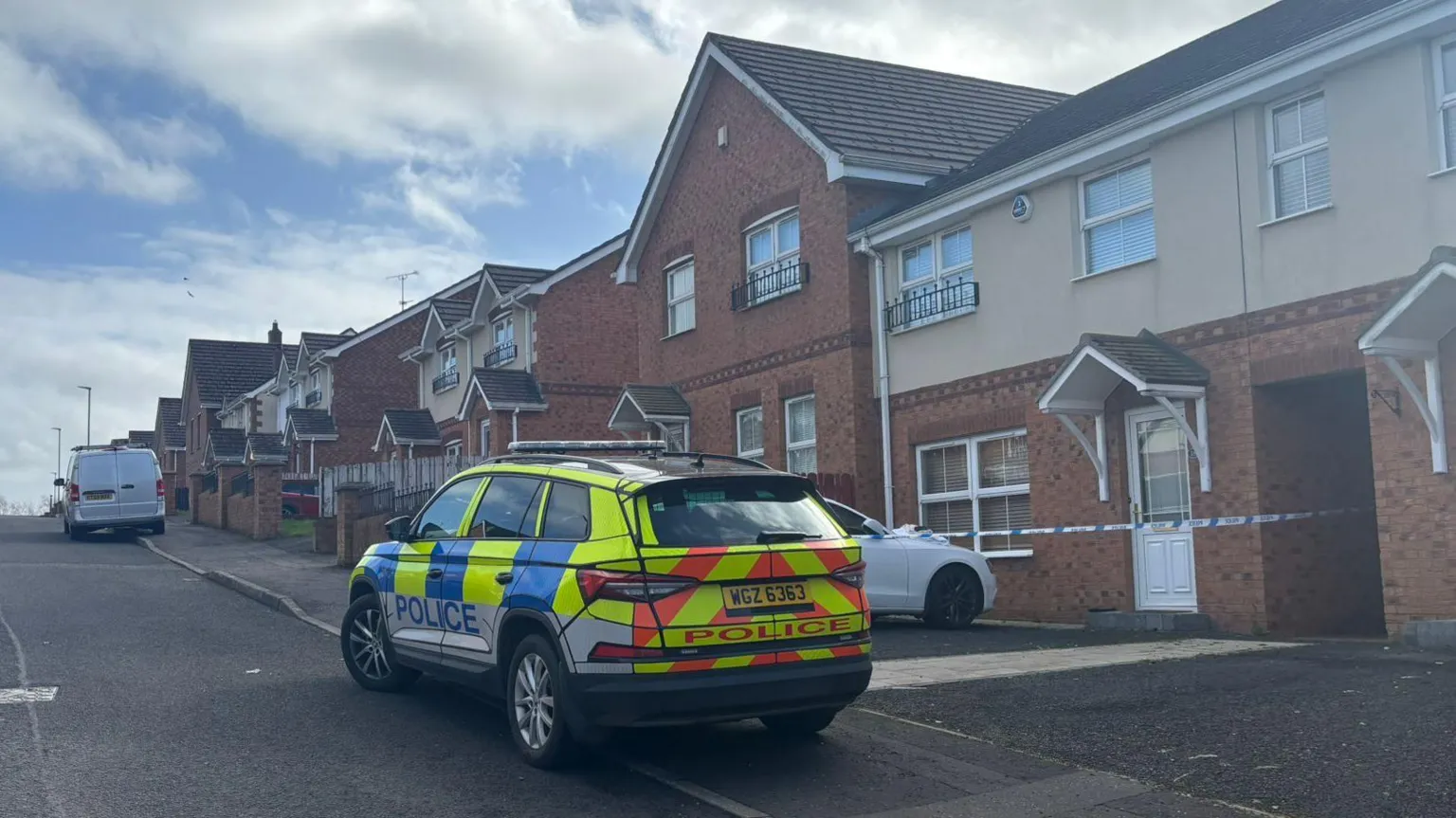 A yellow and orange hi-viz coloured police car sits in front of a row of red-bricked semi detached houses. The houses sit to the right of the frame, with the car to the left. There is blue and white police tape that runs in front of one the houses. A white car is parked in the house's drive way. 