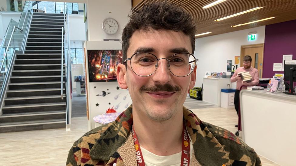 Scott Thomas, dyn ifanc gyda gwallt a mwstash brown a sbectol, mewn cyntedd canolfan