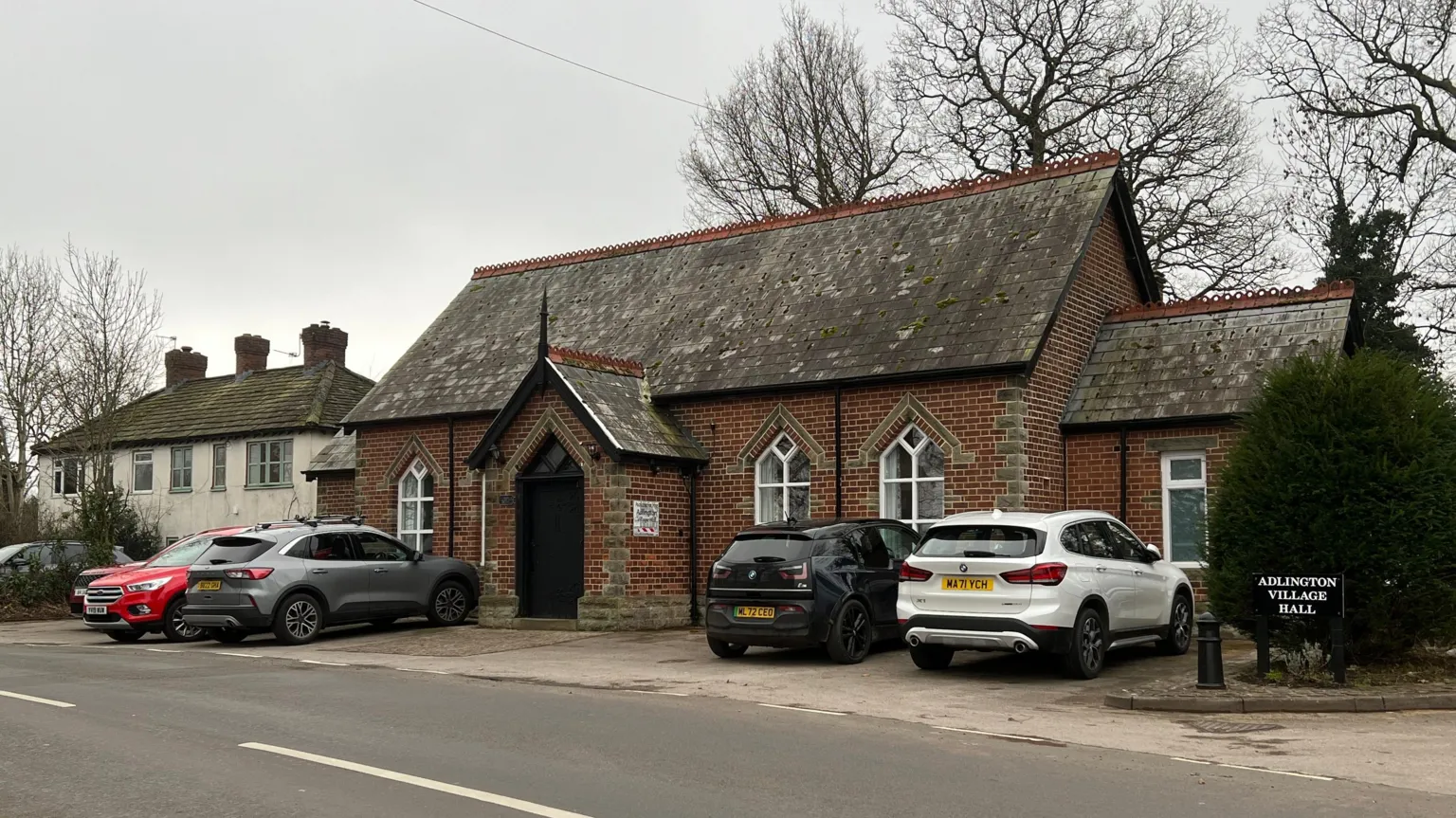 A view of Adlington VIllage Hall. It is an old redbrick building and has five cars parked outside.