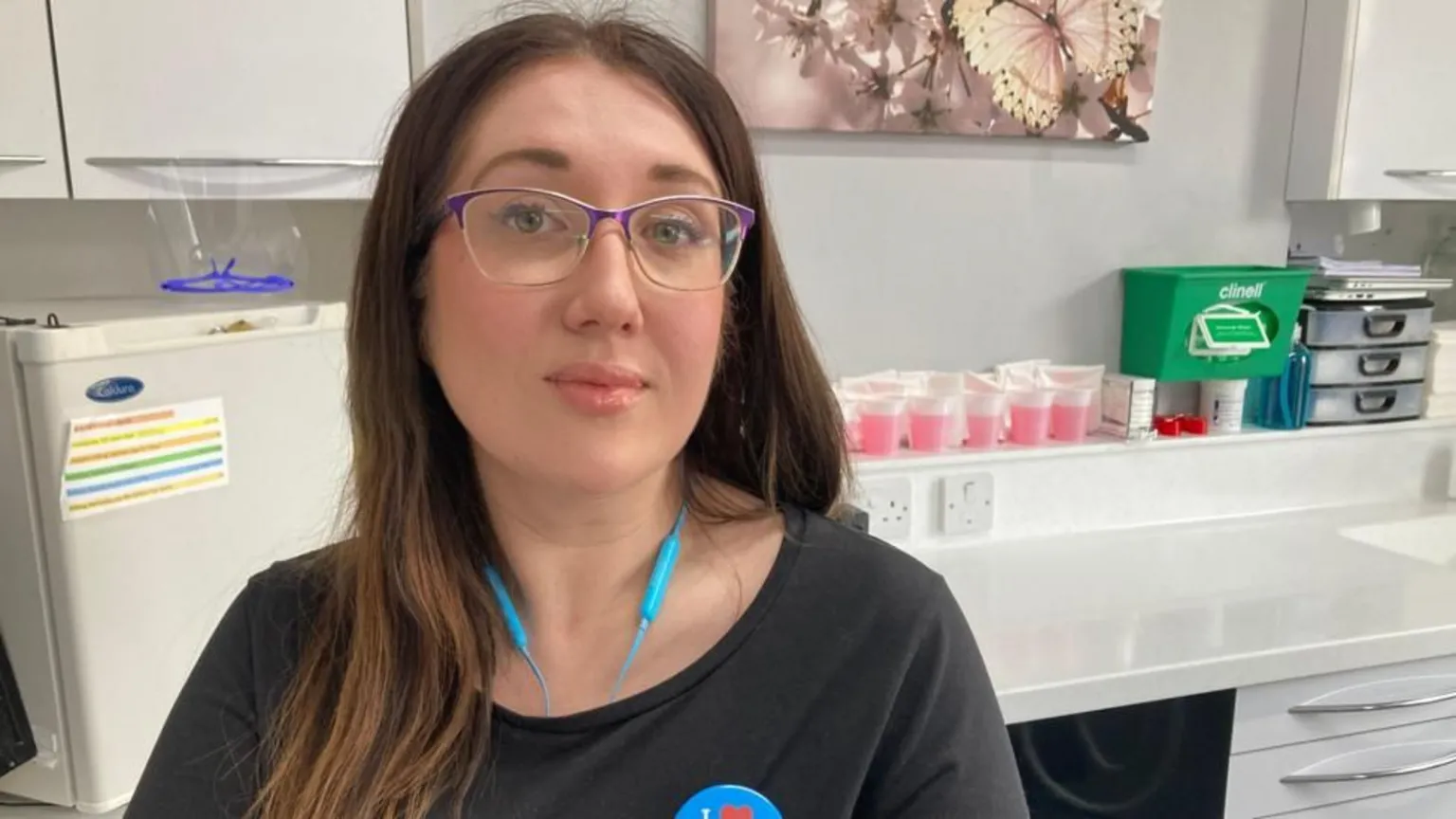 A woman with brown long hair and glasses with a purple edge is looking at the camera. Behind her is a fridge, cups with pink liquid and a green medical box.