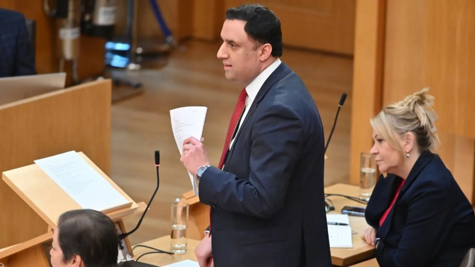 Anas Sarwar, who has short, black hair, stands up at a podium in the Scottish Parliament. He is wearing a dark suit, white shirt and red tie, and holding white papers . 