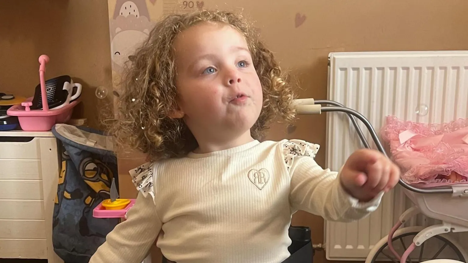 Grayce Pearson has an animated look on her face. She is looking and pointing at someone off camera. She has shoulder-length, curly, blonde hair and is wearing a long-sleeved cream top with frills on the shoulders. There are children's toys, including a pram, behind her.