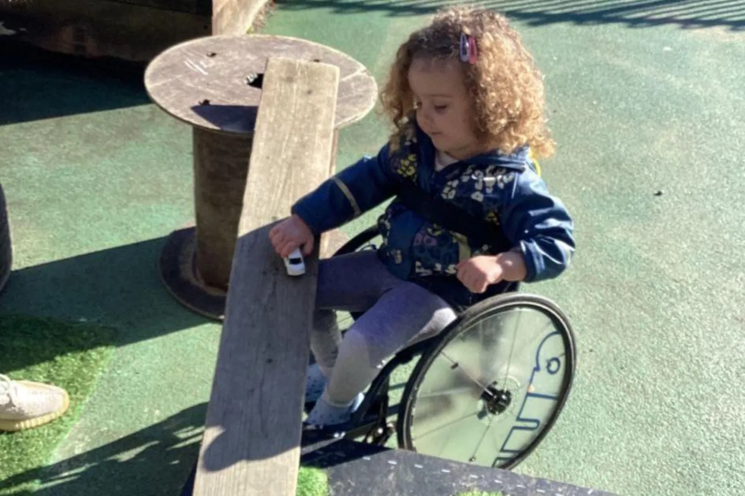 Pearson family Grayce Pearson is sitting in a small wheelchair while playing with a toy car on a wooden plank which is set up like a bridge. Another toy vehicle is on the artificial‑grass-covered section of the platform. Grayce has shoulder-length, curly, blonde hair and is wearing a blue hoodie and grey leggings.