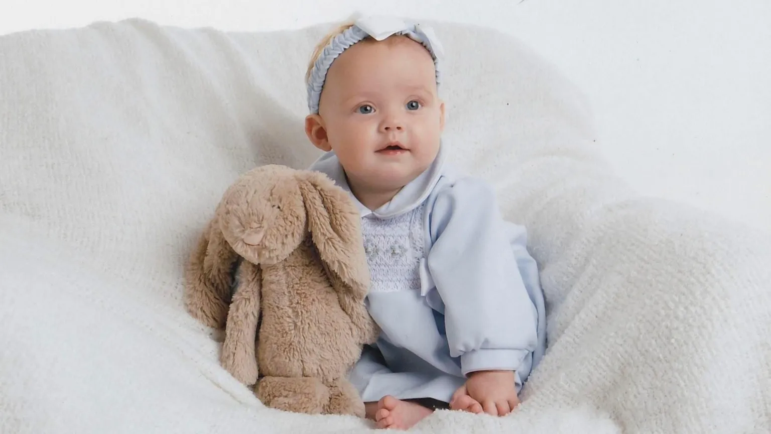 Forever Timeless Photography Professional studio photograph of Grayce Pearson as a baby. She has a half smile and is wearing a baby blue dress and matching hairband with a bow. Her toes are visible and she is sitting with a brown Jellycat rabbit.