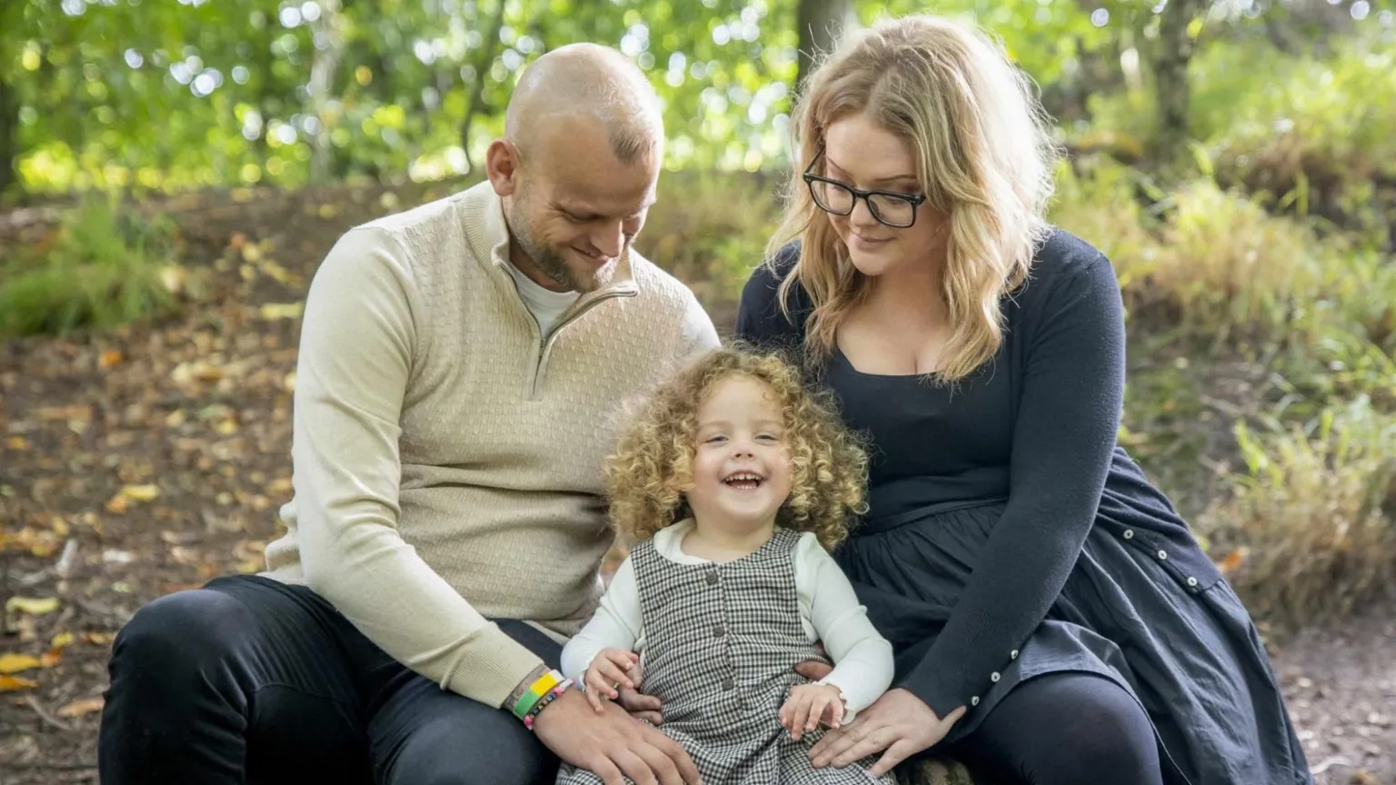 Forever Timeless Photography Tony and Carrie Pearson sitting outside with their daughter Grayce. Grayce is smiling widely at the camera and has shoulder-length, curly, blonde hair. She is wearing a white, long-sleeved top and a checked brown pinafore dress. Carrie is wearing a black top and skirt and has long, blonde hair and black-rimmed glasses. Tony has a shaved/bald head and a beard and is wearing a beige, long-sleeved top. There are trees and grass behind them.