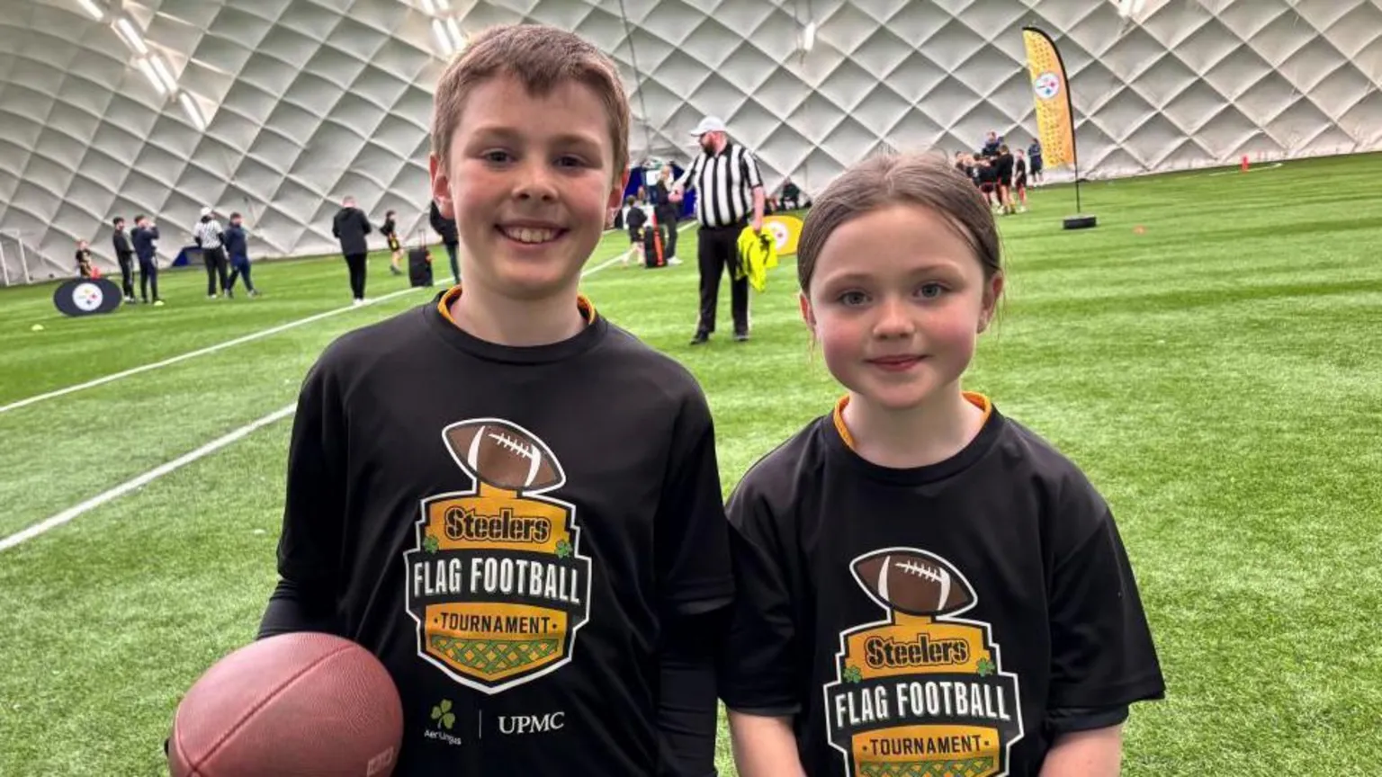 A young boy and girl standing side by side. They are booth wearing black tops with 'Steelers flag football tournament' on it. Behind them is the pitch. The boy has short brown hair and the girl has long brown hair tied back in a ponytail.