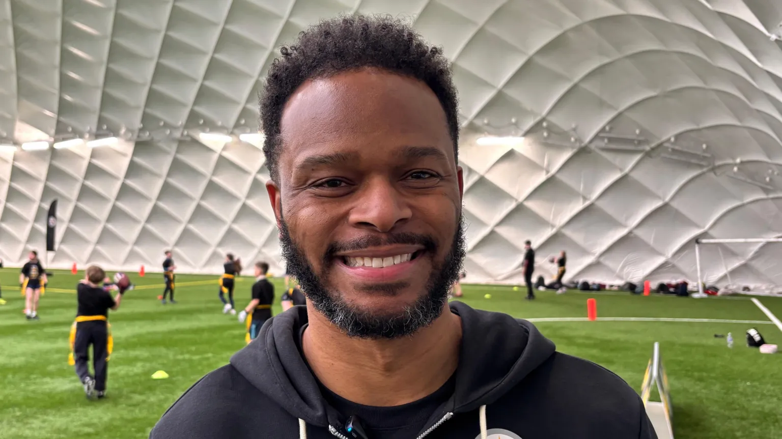 A man with short black hair and facial hair smiling at the camera as he stands in front of kids playing sports. He is wearing a black jacket.