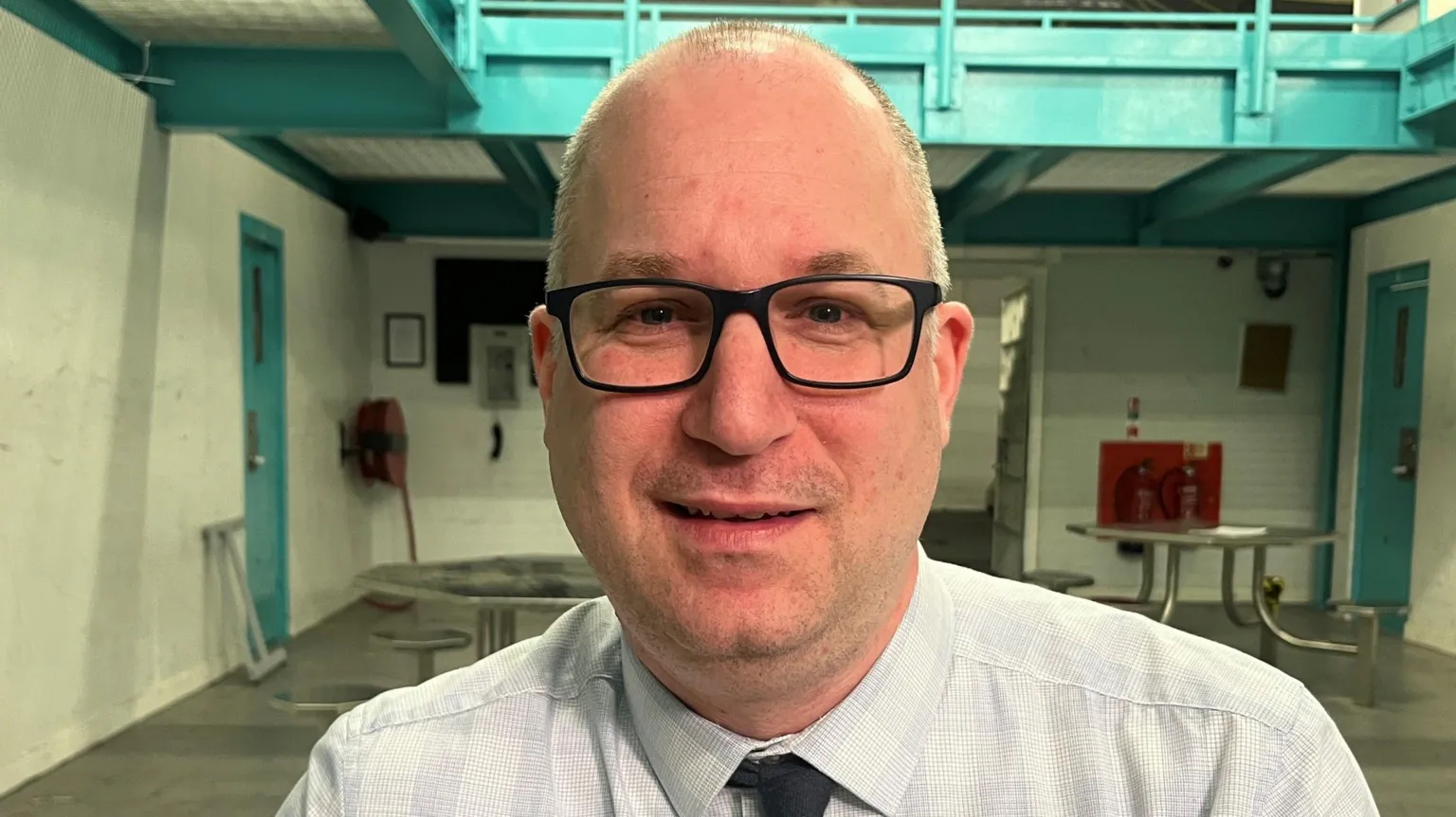 Dr Frank Slokan looking straight at the camera and wearing a white shirt with a dark tie. He is also wearing black-rimmed glasses 