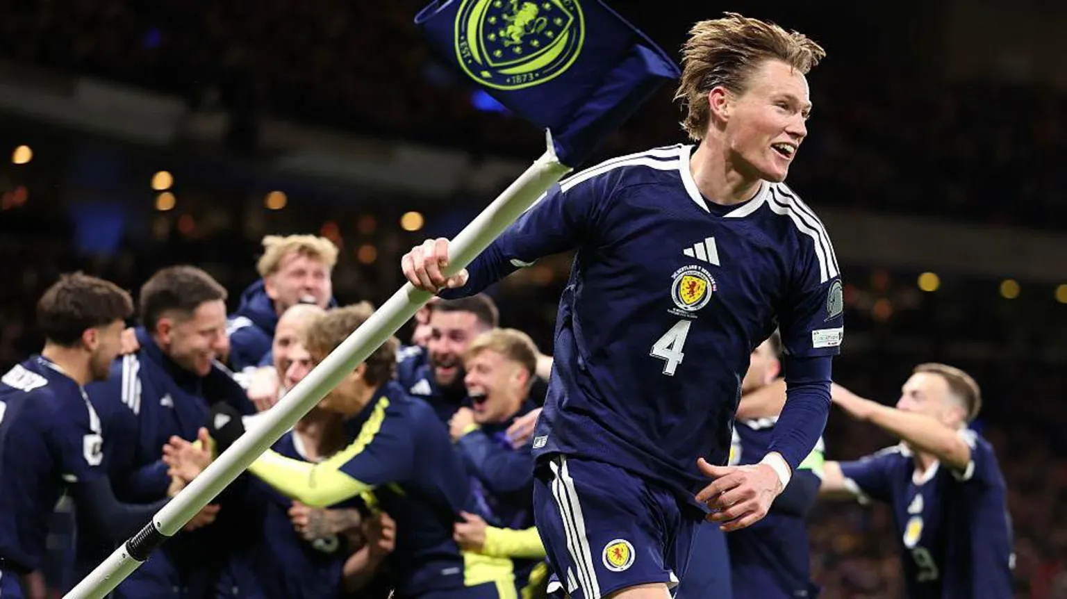  Scott McTominay celebrating with the corner flag. He is wearing a navy blue Scotland shirt and shorts.