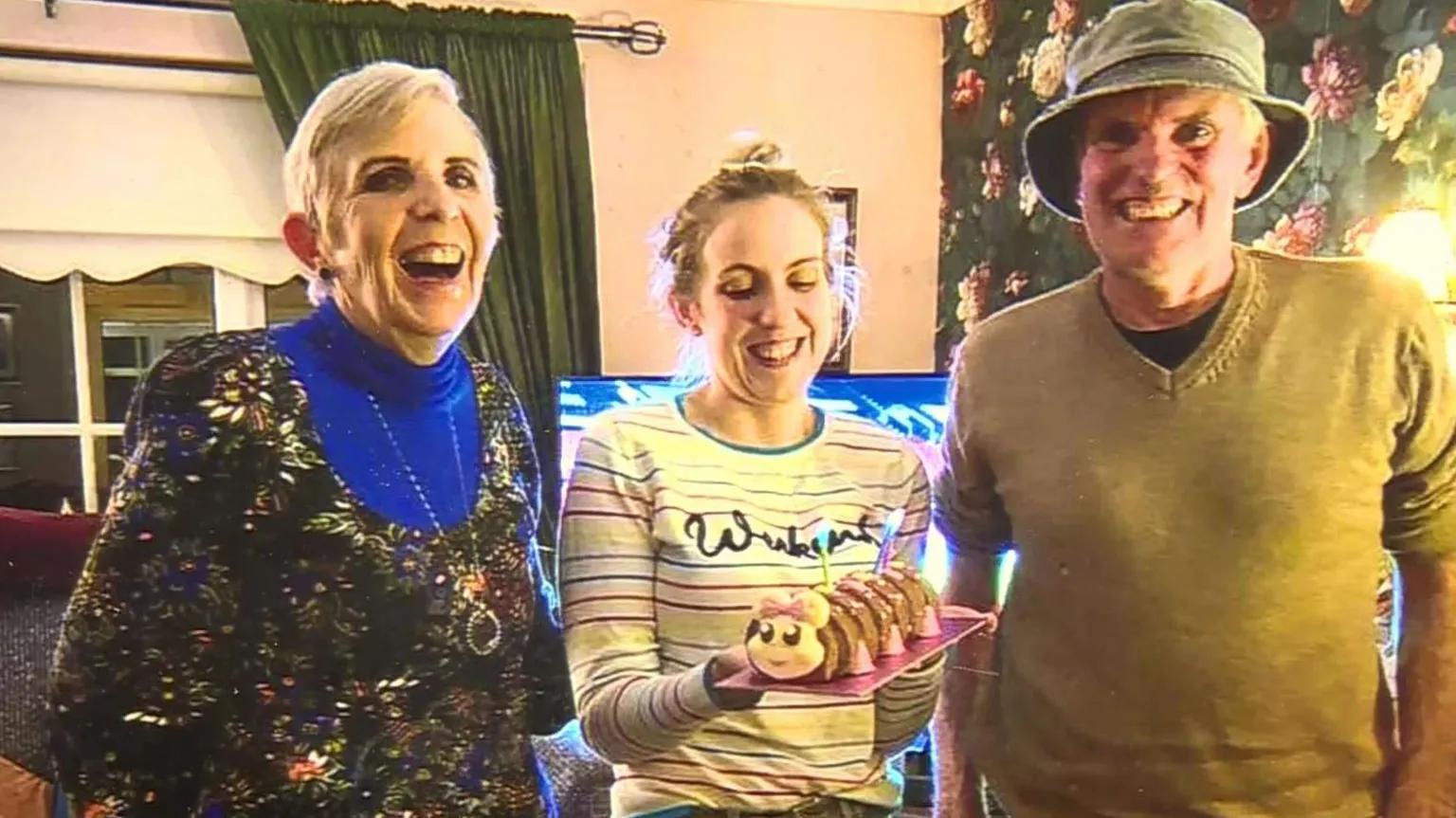 A woman and man stand either side of a young woman. The older woman is laughing, wearing a blue jumper and sparkly top. The older man is wearing a green hat, and a v-neck mustard jumper. He is also smiling. Natalie in the middle, is also smiling She is holding a caterpillar birthday cake with candles on it. She is wearing a pale yellow striped top. Dark curtains and a window are visible in the background.