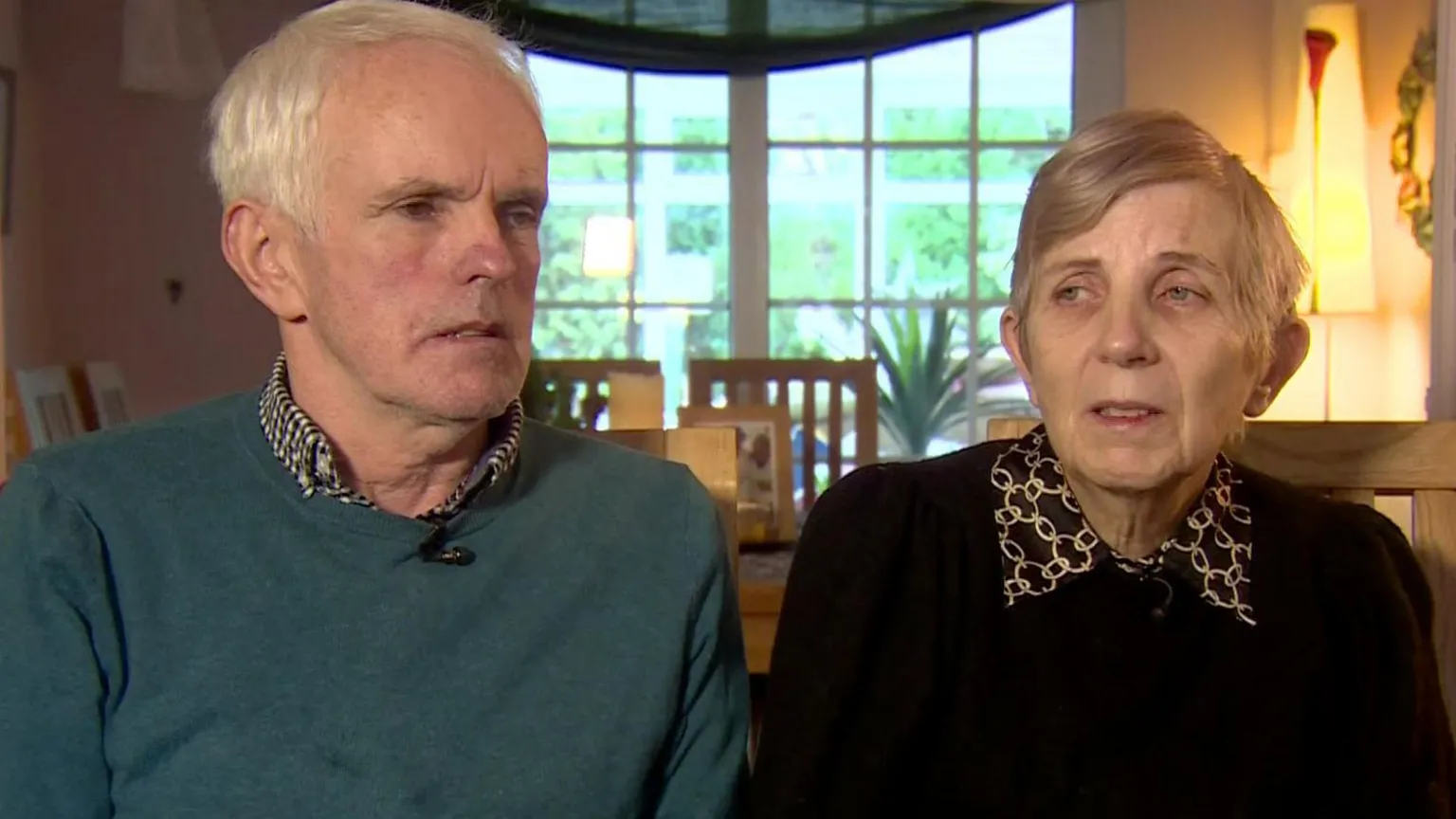 A man and a woman sit beside each other during a TV interview. The man, Noel McNally, has short white hair and is wearing a dark green jumper over a grey patterned shirt. The woman, Bernie McNally, has short blonde hair and is wearing a black jumper over a black and gold chain patterned blouse. There is a window and a dining room table in the background. 