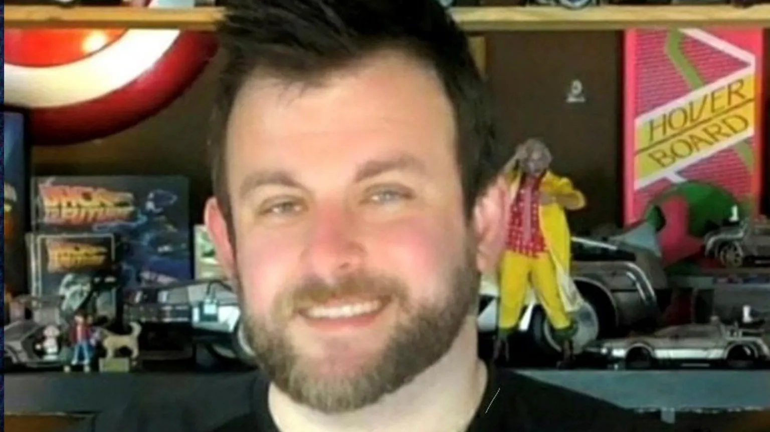 Stephen McCullagh, a man with short dark hair and a beard, smiles at the camera. He us wearing a black crew-neck top or t-shirt. He is standing in front of a shelf containing retro toys including toy DeLorean cars and figures from the film Back to the Future. There is also a sign for a hover board. 