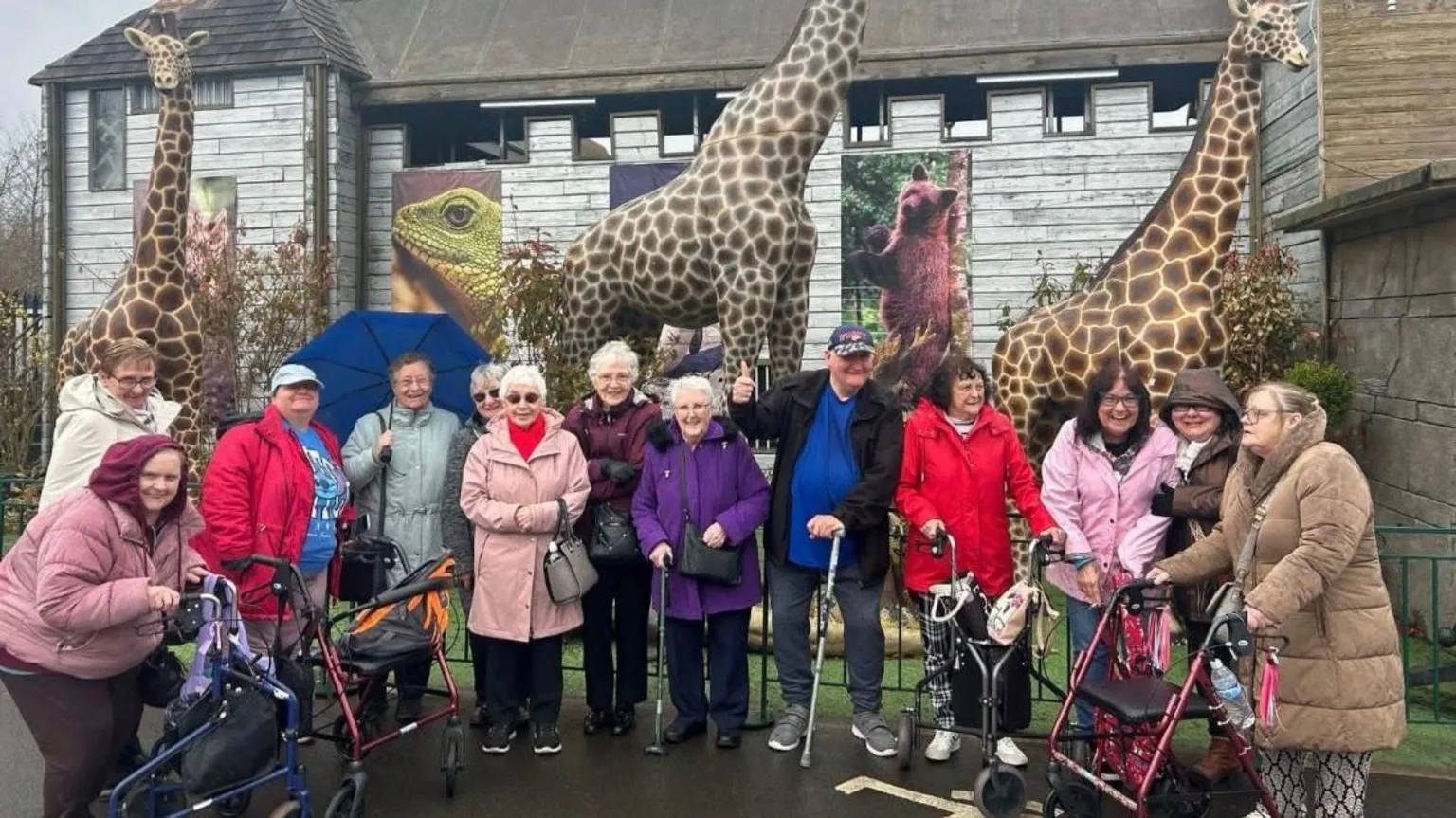 CACE A group of elderly people with walking aids, standing at a zoo entrance with life-size models of giraffes behind them as well as large photographs of a bear and a lizard