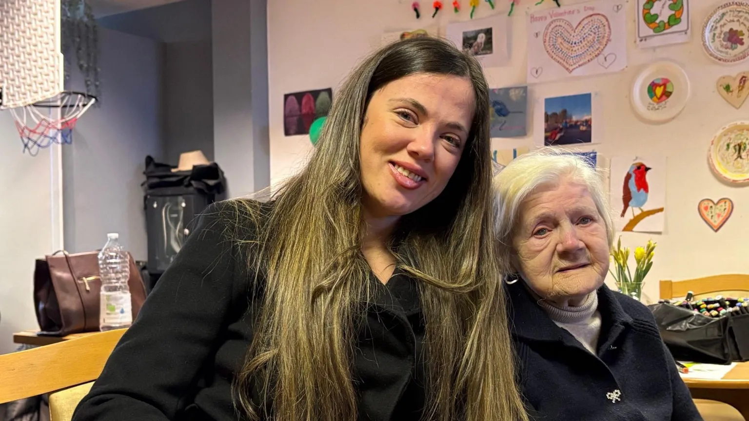 Brooke, who is in her mid 30s has long, brown hair. She is smiling at the camera and sitting next to her grandmother who has short, grey hair and is looking at the camera. She is wearing a grey jumper and a dark jacket with a silver brooch. Brooke is wearing a dark shirt or jacket. There are home-made drawings on the wall behind them. 