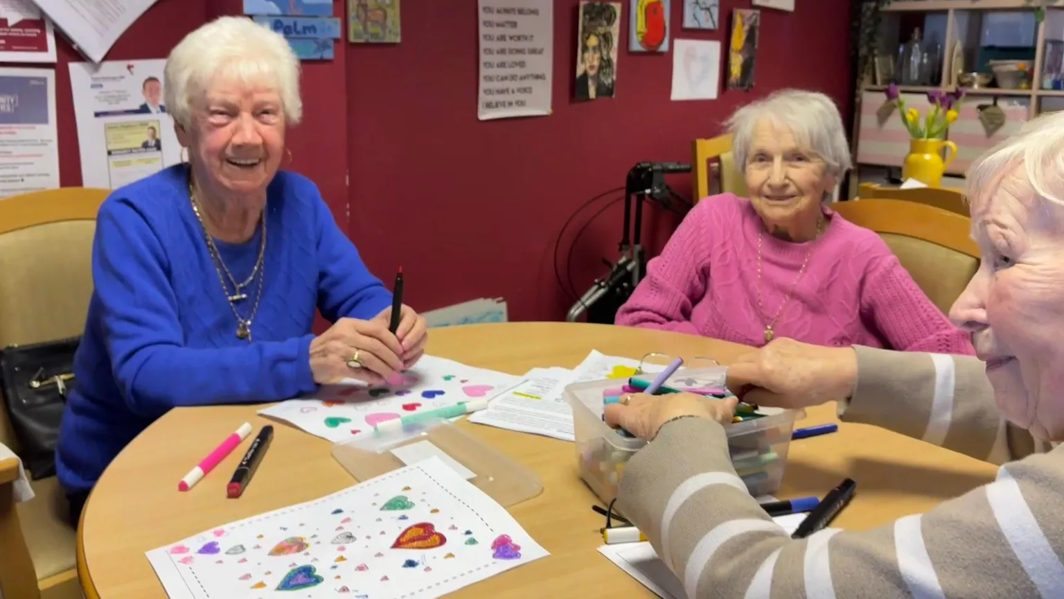 Three pensioners, Alice, Mary and Margaret sitting at a table with felt-tip pens and colouring sheets. They all have short, white or grey hair. Alice is wearing a blue jumper and gold necklaces, Mary is wearing a pink jumper and Margaret is wearing a brown and white striped jumper.
