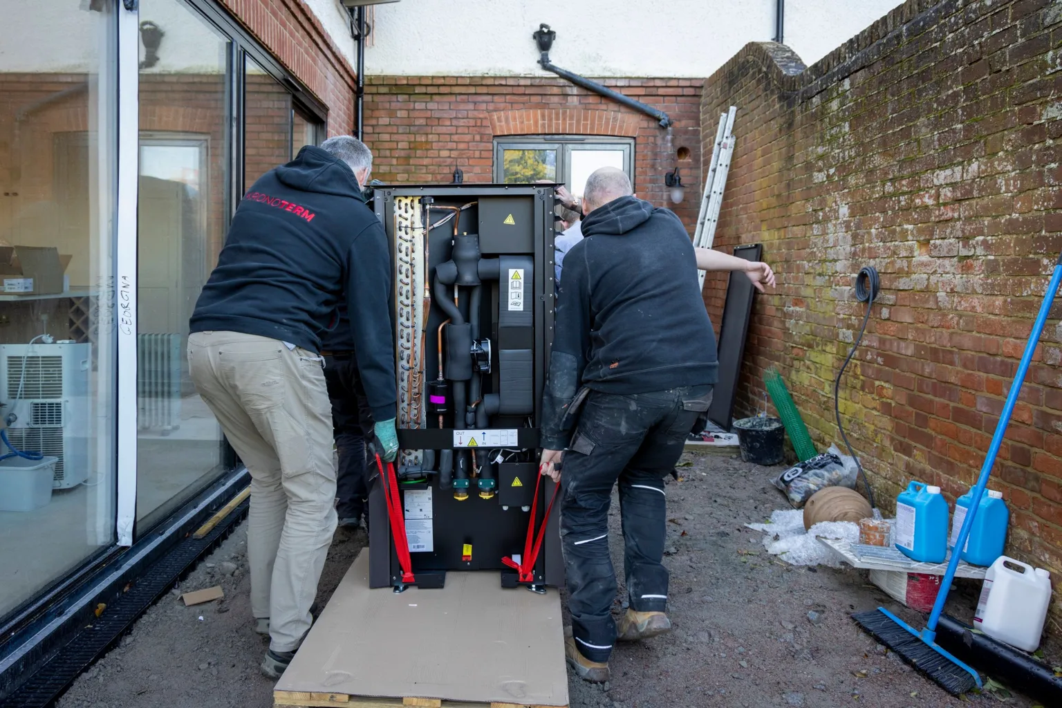 Andrew Aitchison/ Three men with their backs to the camera are shown lifting a black heat pump on a trolley towards the back of the house. The men wear dark work clothes and boots. The house is red brick with a long glass door to the left and the property boundary wall to the right. Against the wall sits work tools and a blue broom. 