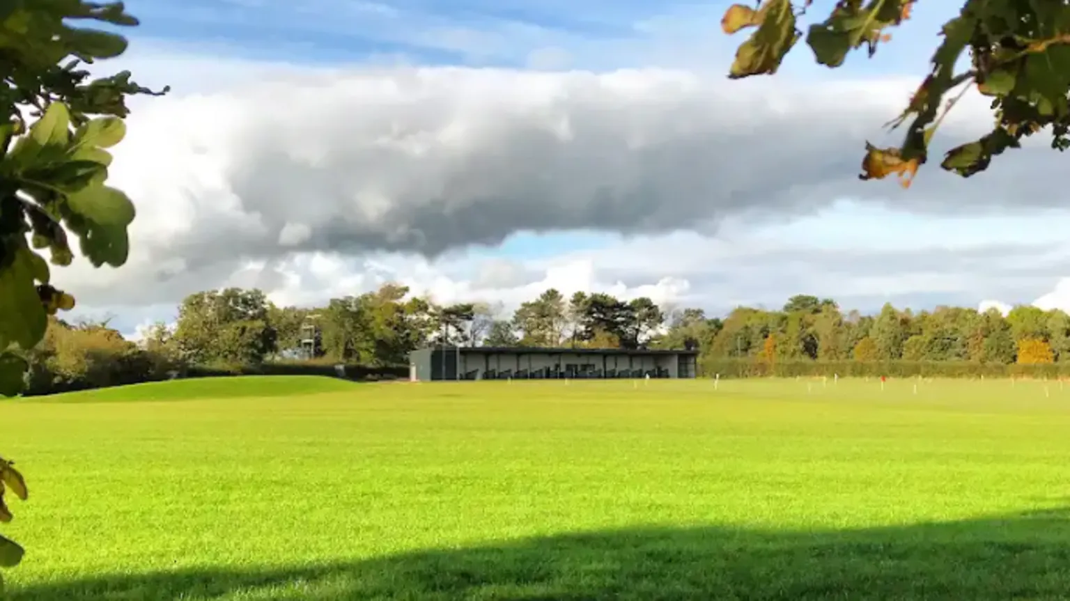 Need Golf A large green field, with sun shining on the grass. There is a long building made of a number of bays across the back of the field