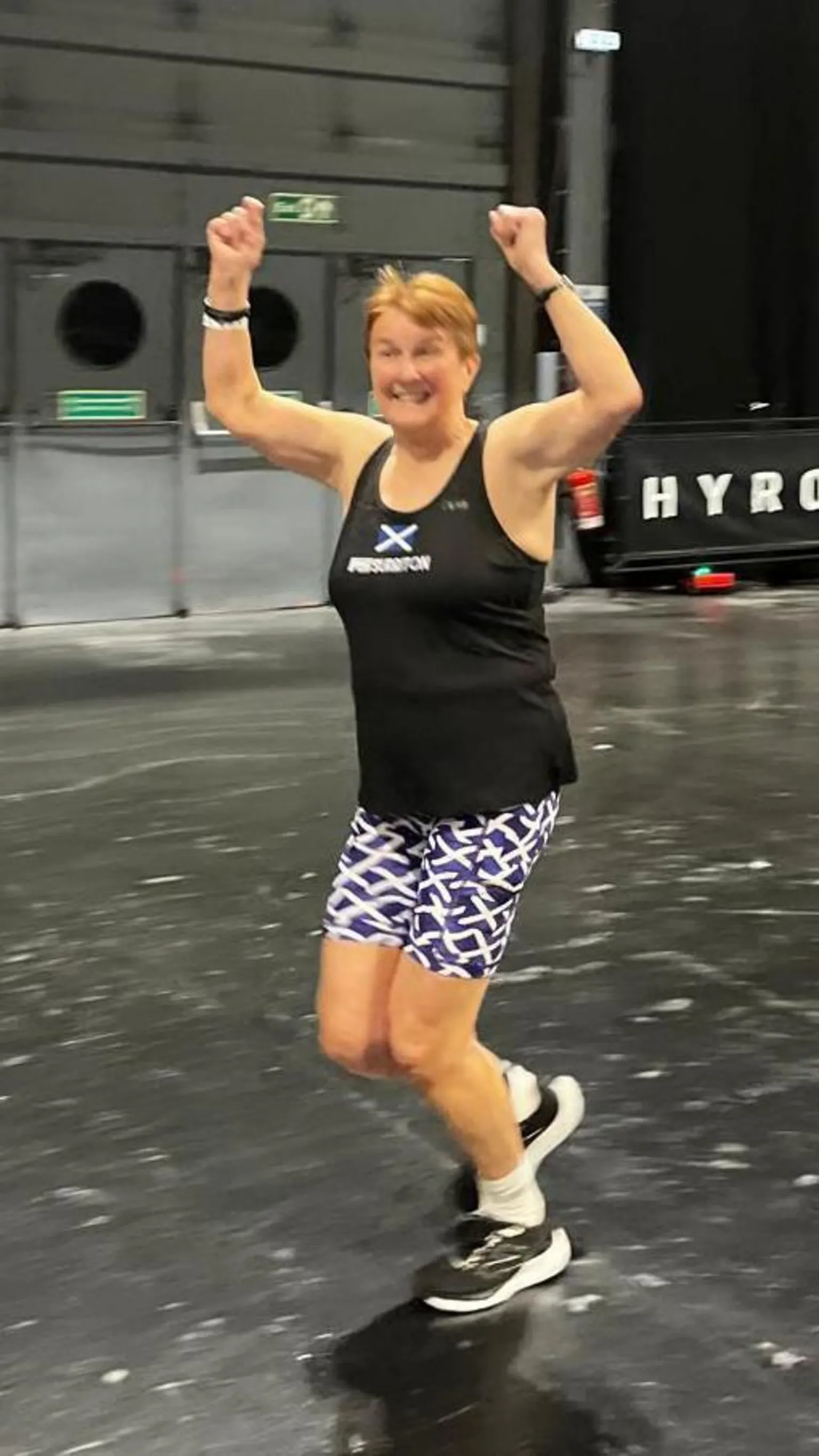 Stuart Amory Carole Munro running during a Hyrox event. She has both her arms in the air and is wearing a black vest top and Saltire-flag print shorts.