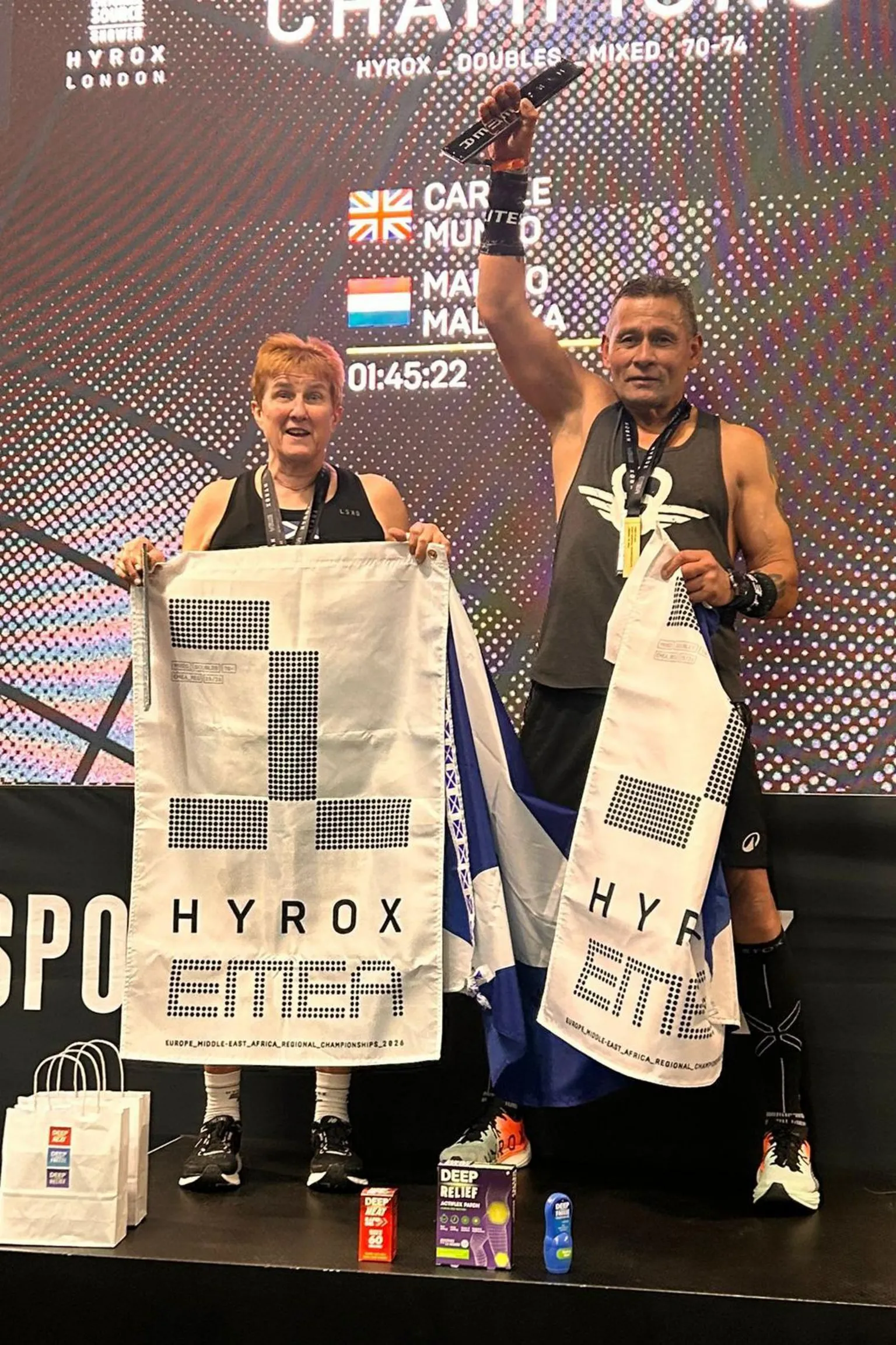 Stuart Amory Carole Munro and Jim Malinka stand on the podium holding white and black Hyrox flags.