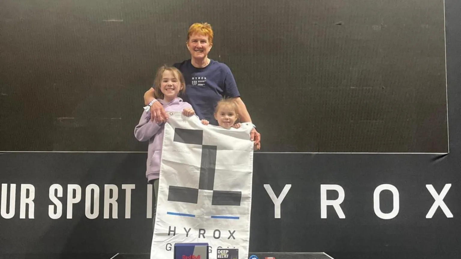 Stuart Amory Carole Munro and her two granddaughters aged eight and four standing on a podium holding a white and black Hyrox flag. 