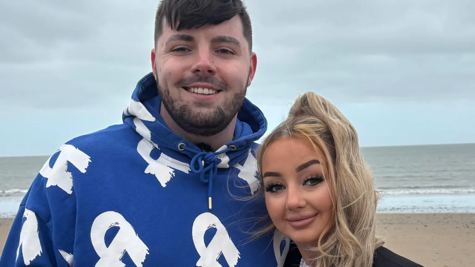 Ruben and partner Amber smiling at the camera on Abersoch beach with the sea behind them. Amber has a long blond ponytail. Ruben is wearing a blue hoodie