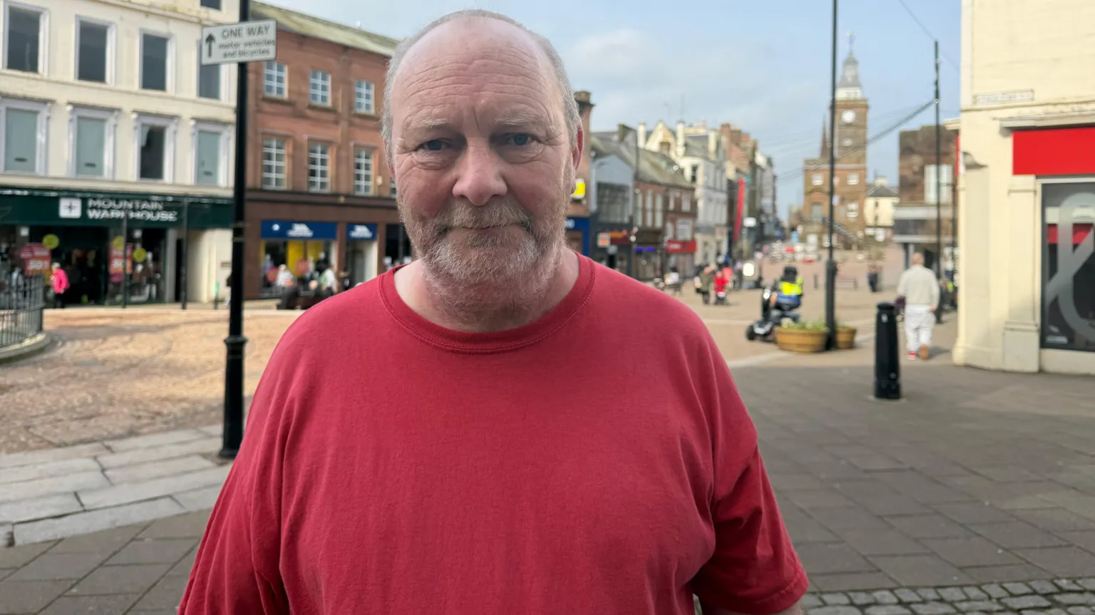 Man in red t-shirt standing on Dumfries High Street with shops and the Midsteeple in the distant background.