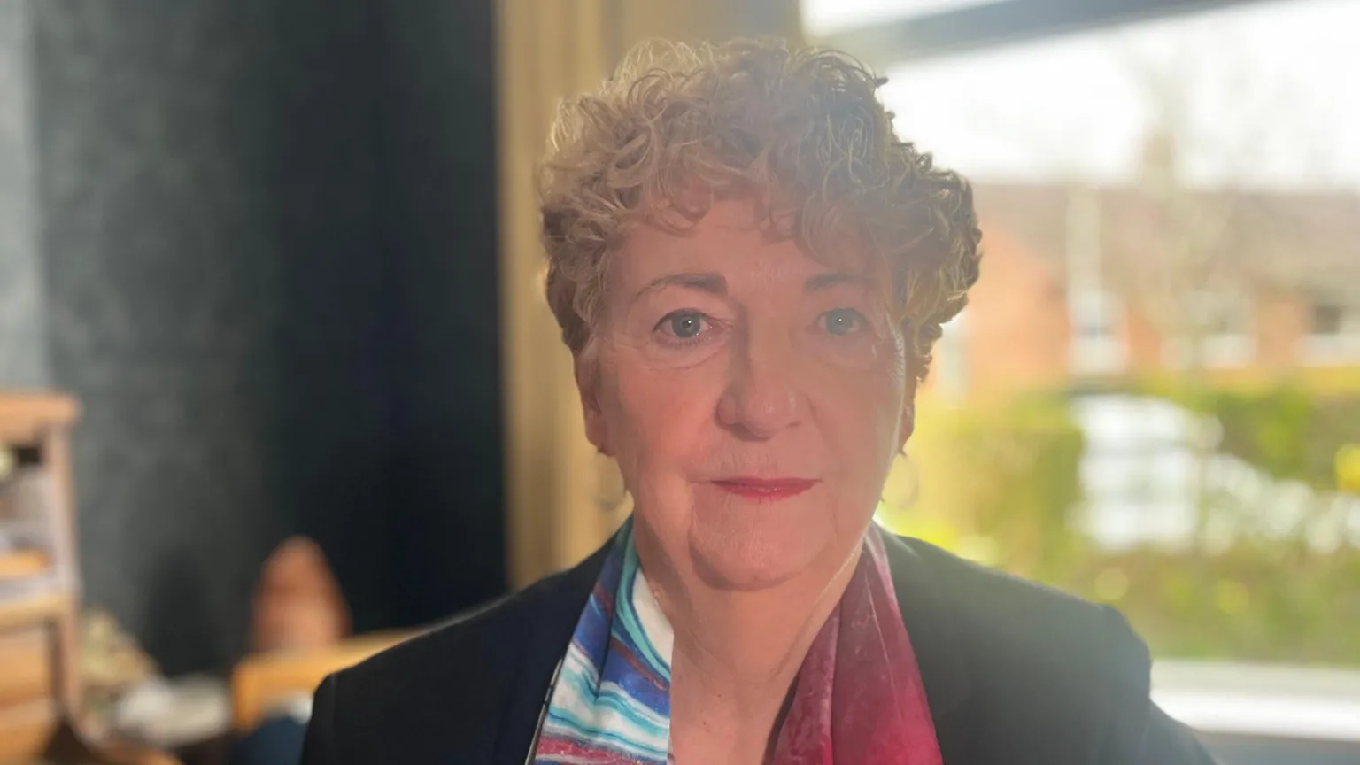 A woman with short, curly blonde hair looks into the camera. She is wearing a dark jacket and a multi-coloured scarf. A window is in the background.