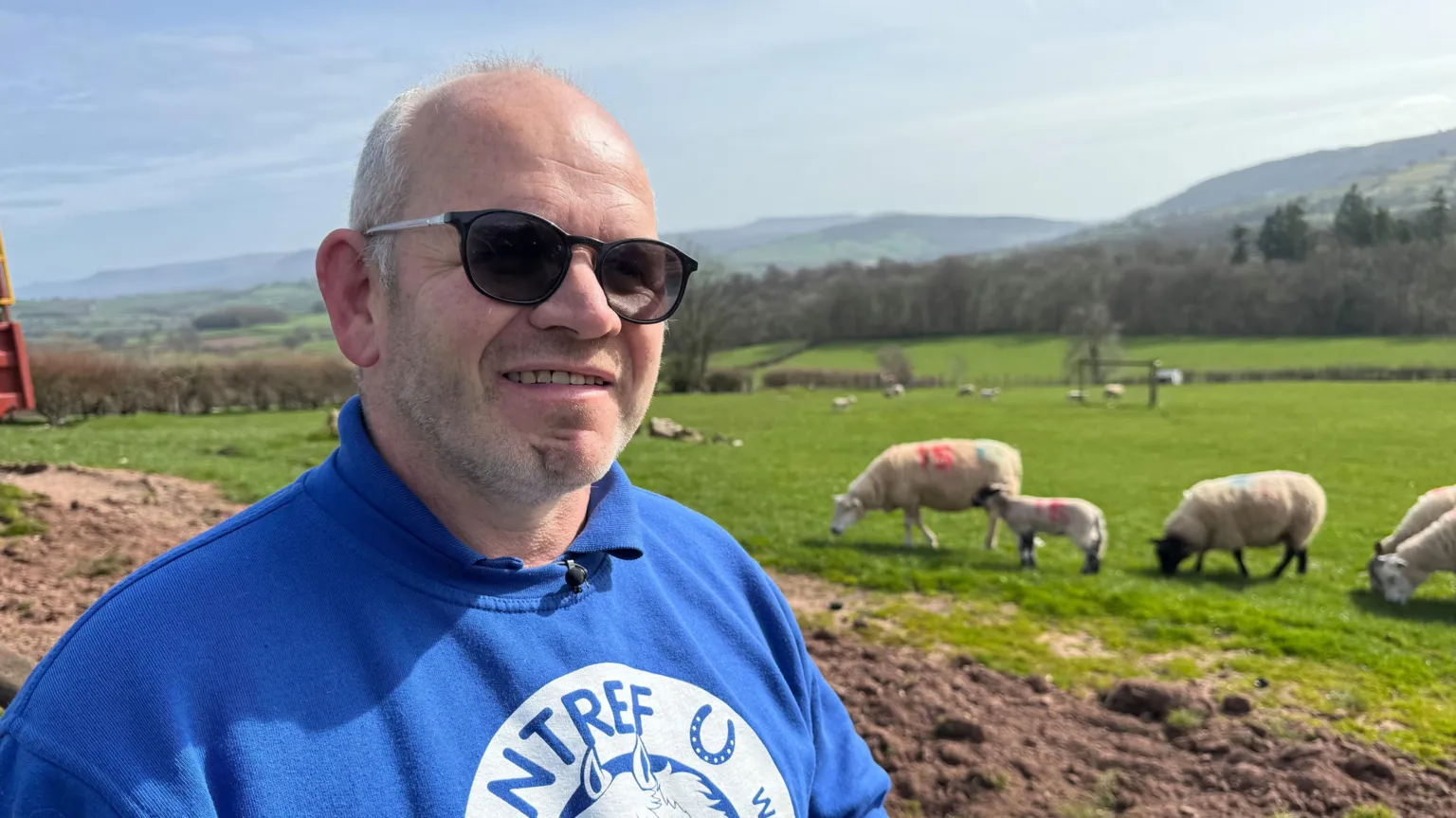 A farmer standing in a blue and white branded polo neck top and jumper with Cantref Farm written on it. He is looking towards the camera on a sunny day as some of his sheep graze in a field behind him.